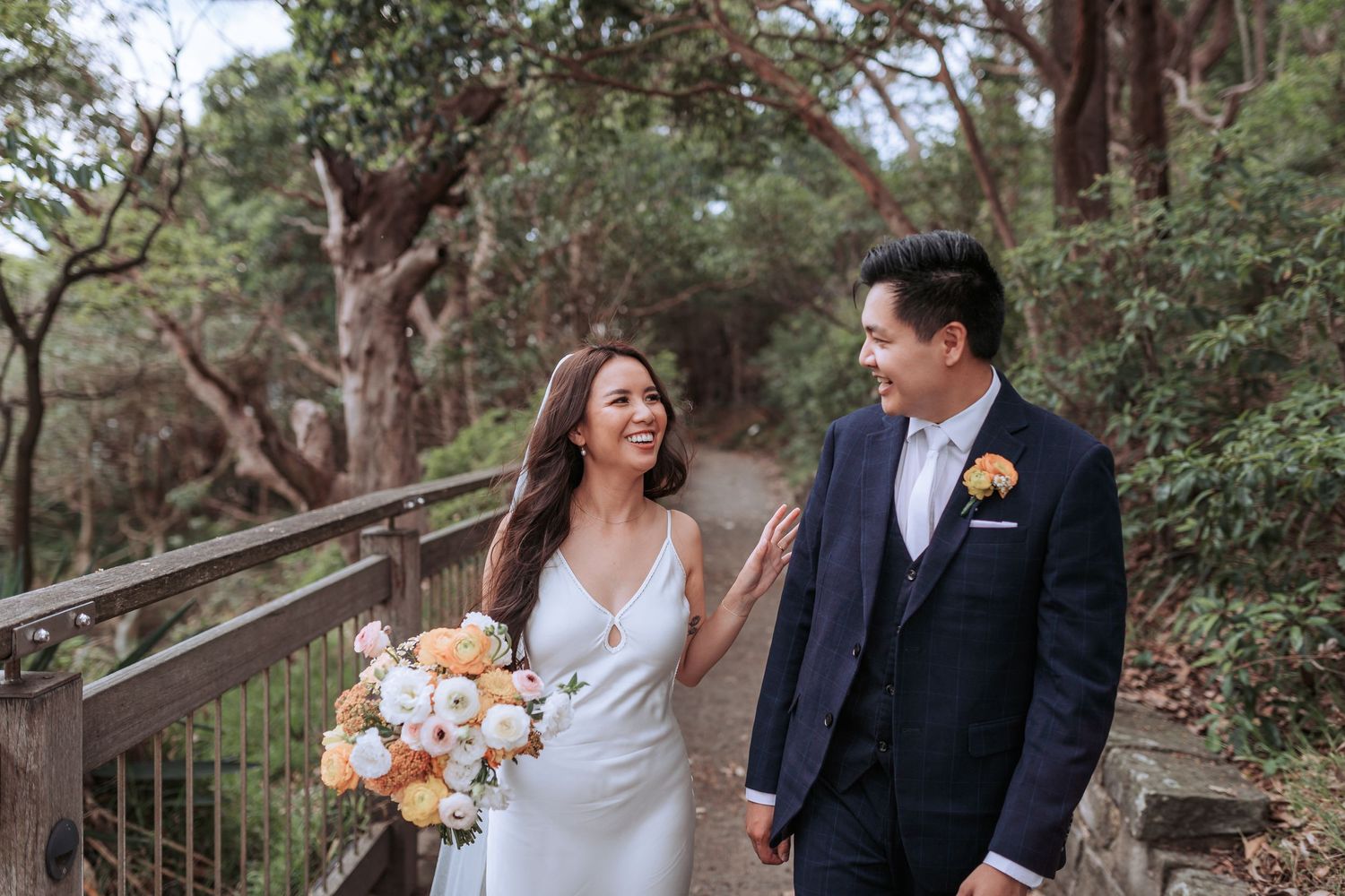 Wedding couple shares joyful laugh while walking on wooden boardwalk path through lush forest setting.