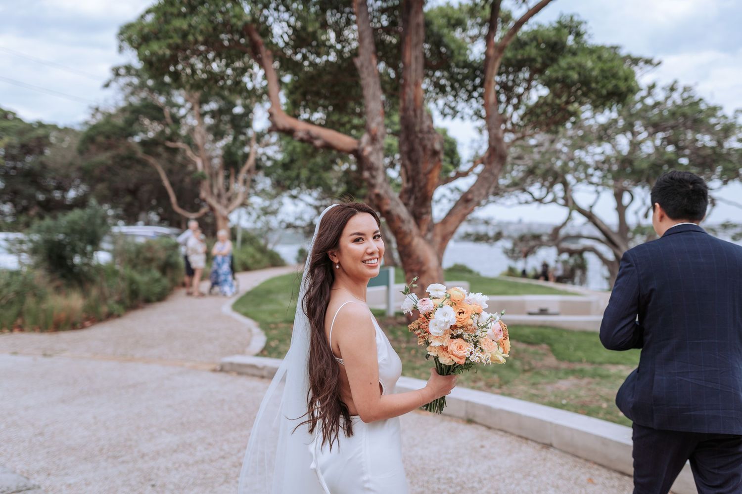 A bride holds a peach and white floral bouquet on a path with eucalyptus trees in the background.