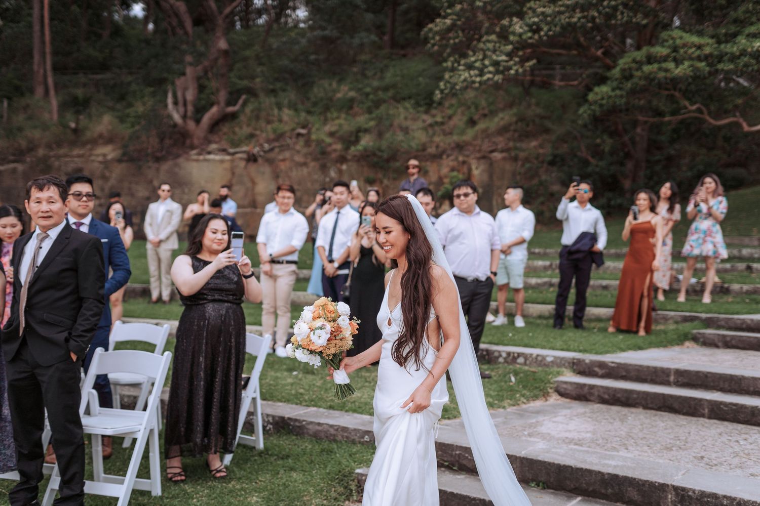 A wedding celebration in a garden setting with guests watching as the bride holds a bouquet of flowers.