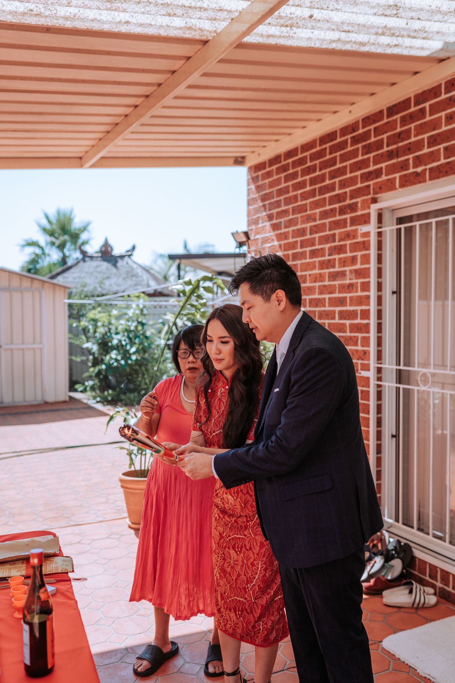Three people gather on a covered patio with brick walls, sharing a moment while looking at something together.