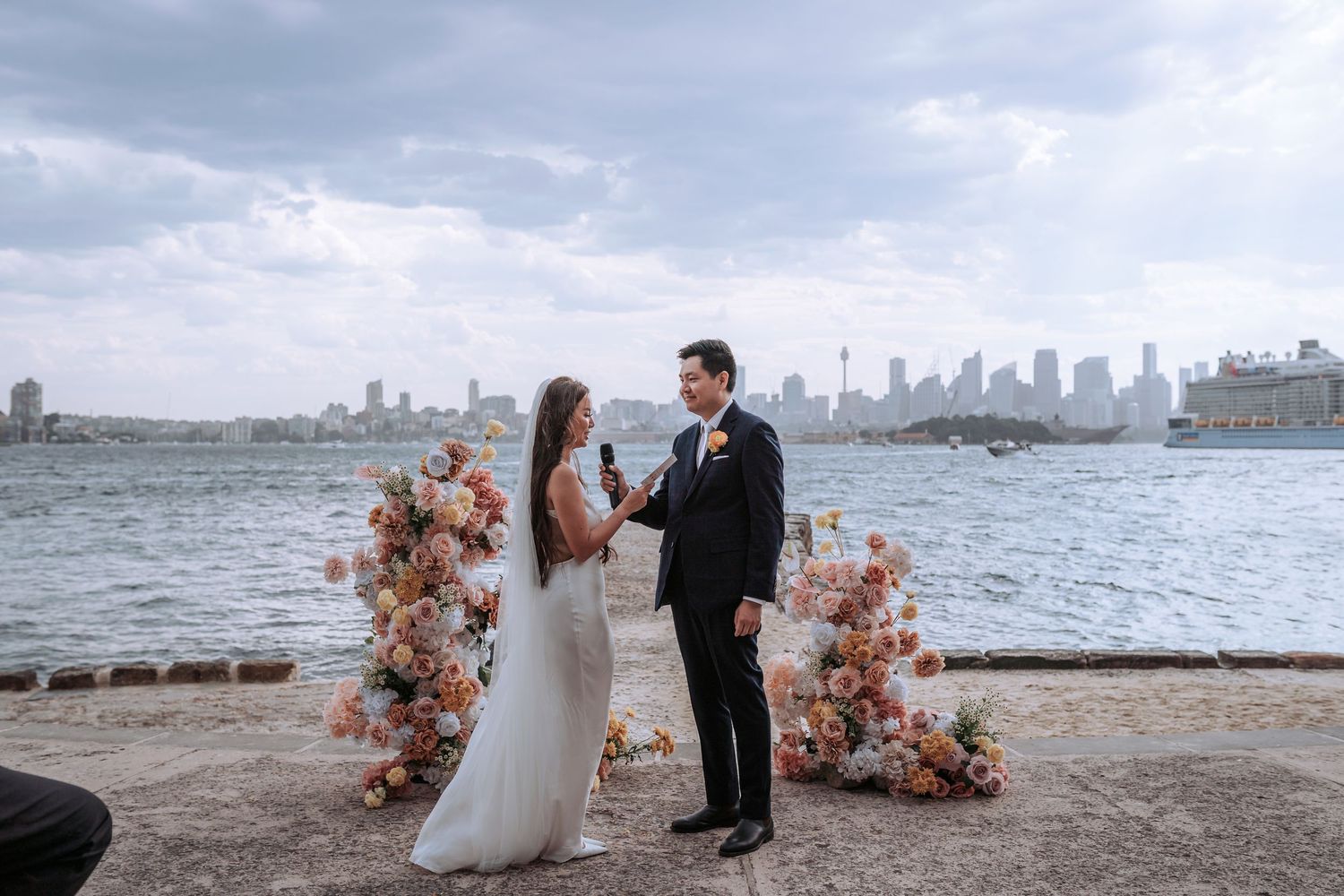 A waterfront wedding ceremony with floral arrangements and city skyline in the background.