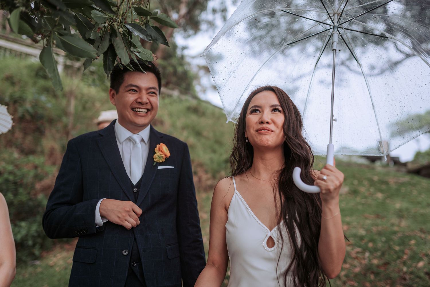 A delighted couple shares a laugh while walking outdoors on their wedding day with the bride holding a white umbrella.