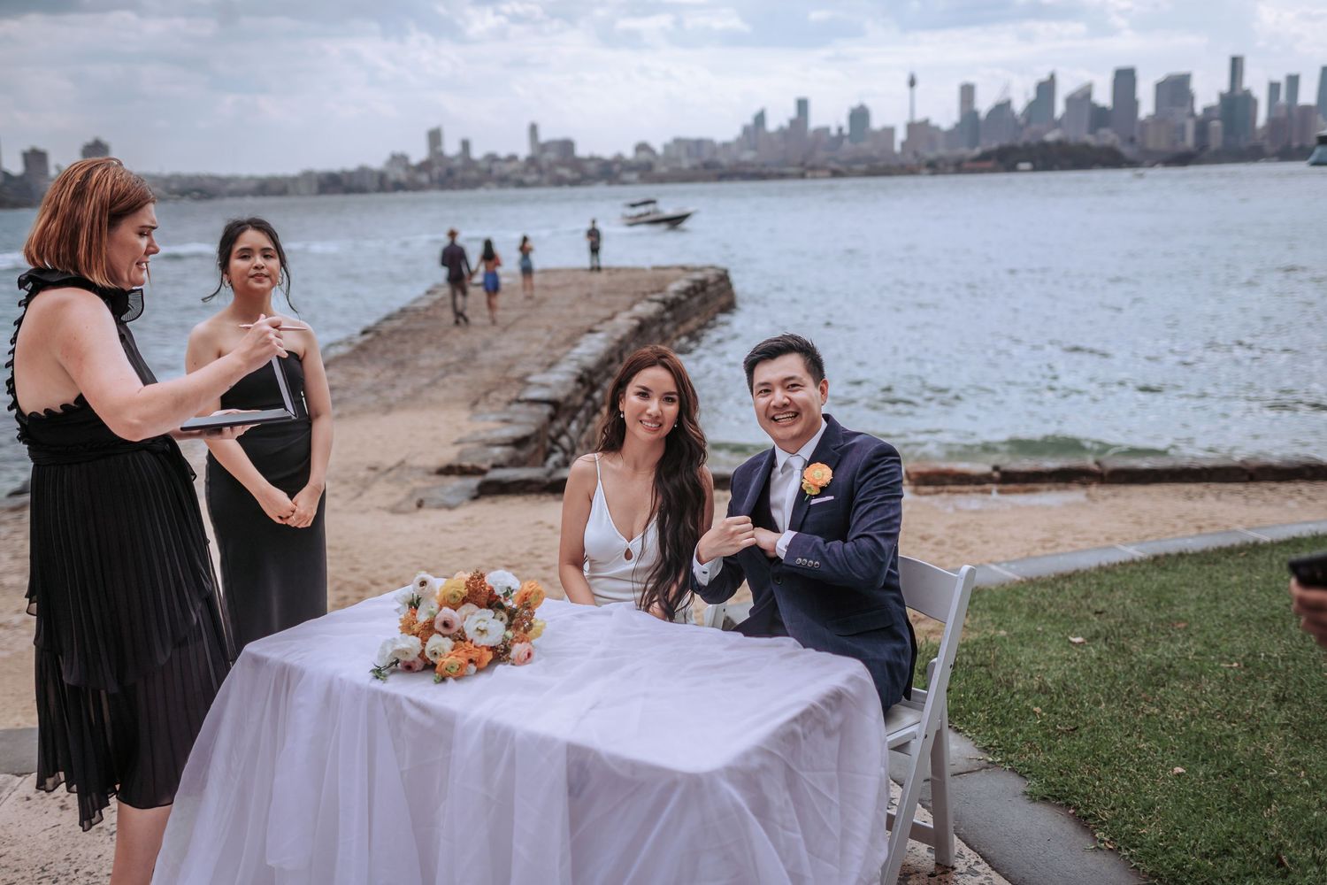 A wedding couple sits at a signing table by the waterfront with the city skyline visible across the harbor.