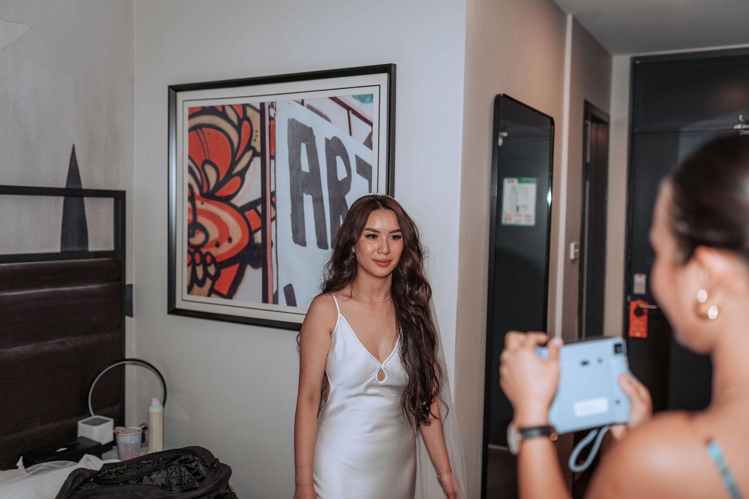 A person in a white dress stands in a room while being photographed with artwork on the wall behind them.
