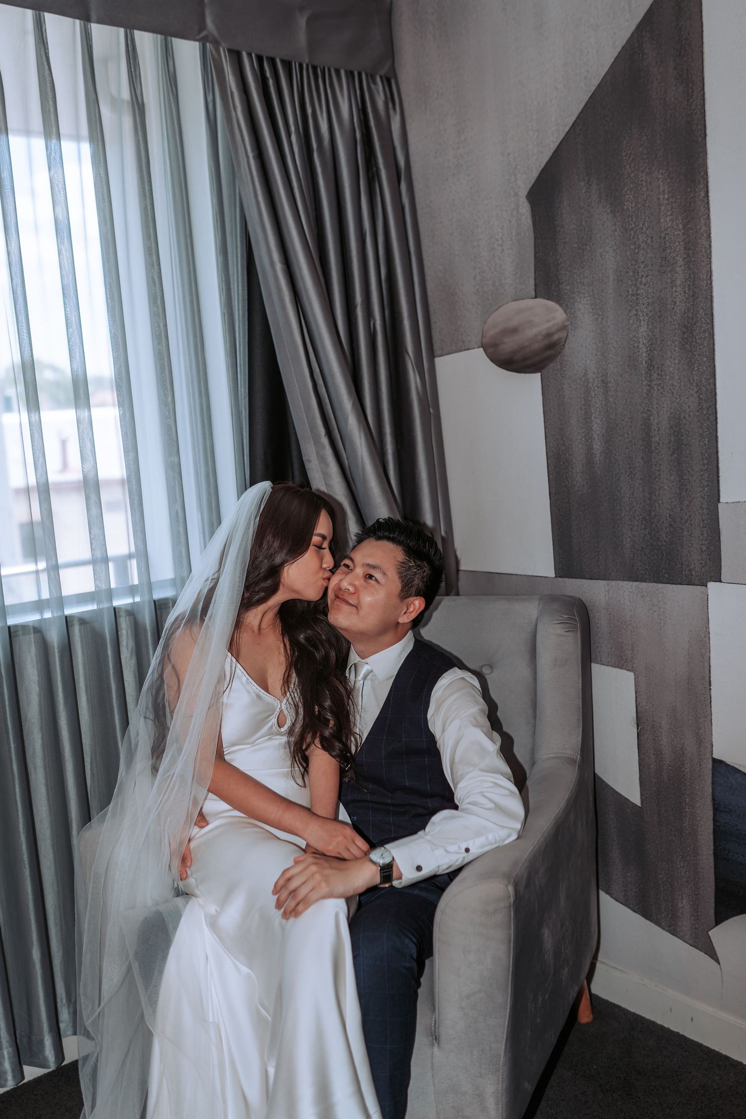 A romantic moment between newlyweds sitting together on a gray couch with elegant curtains.