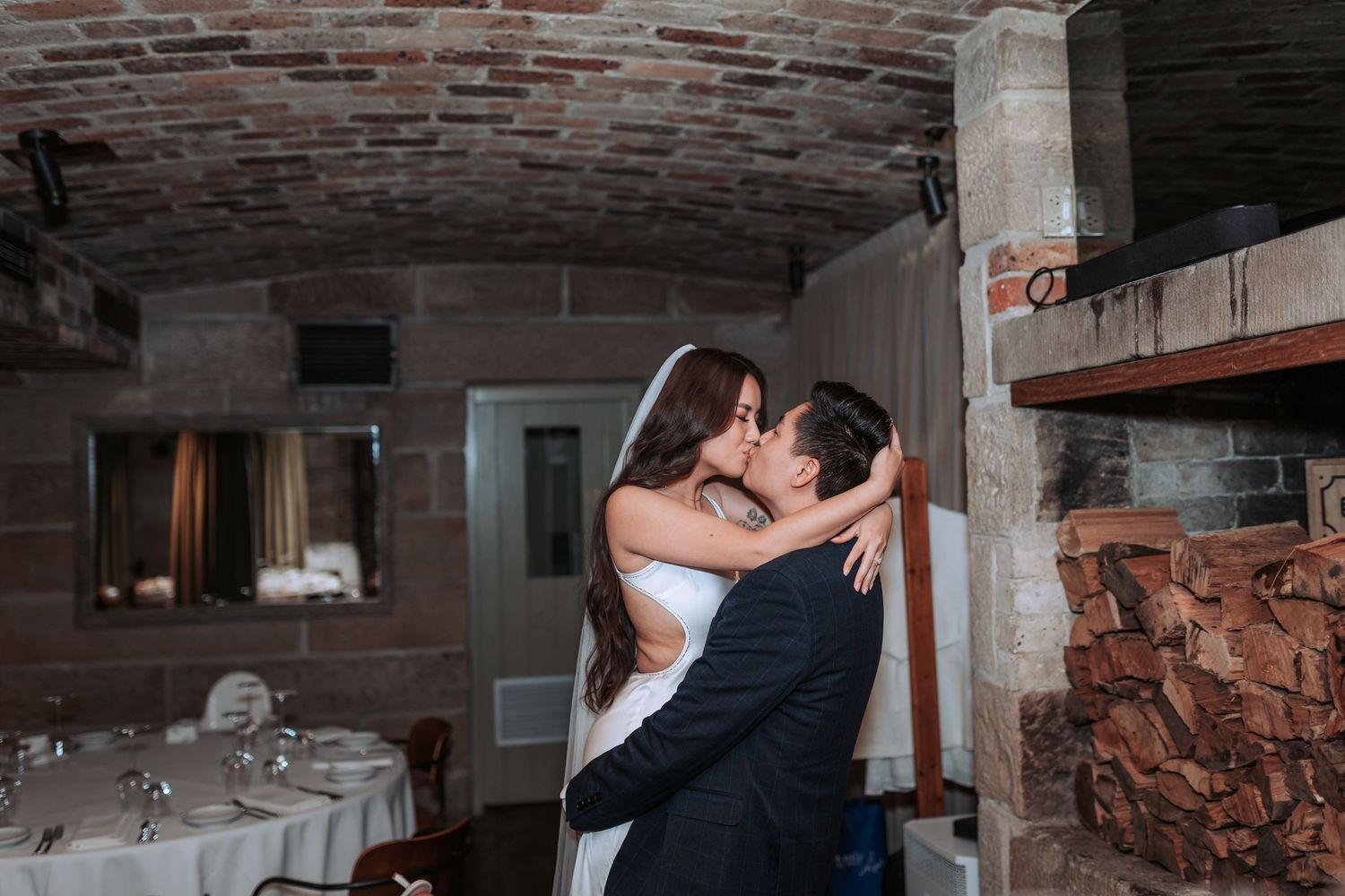 A romantic couple shares an intimate kiss in a rustic brick cellar venue with set dining tables in the background.