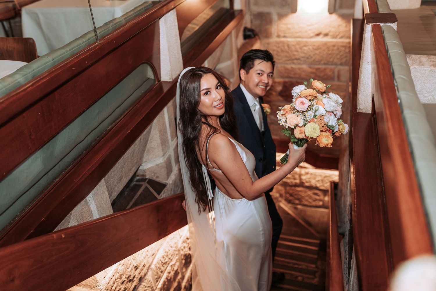 A couple sits together in wooden church pews during their wedding ceremony holding a peach and white floral bouquet.