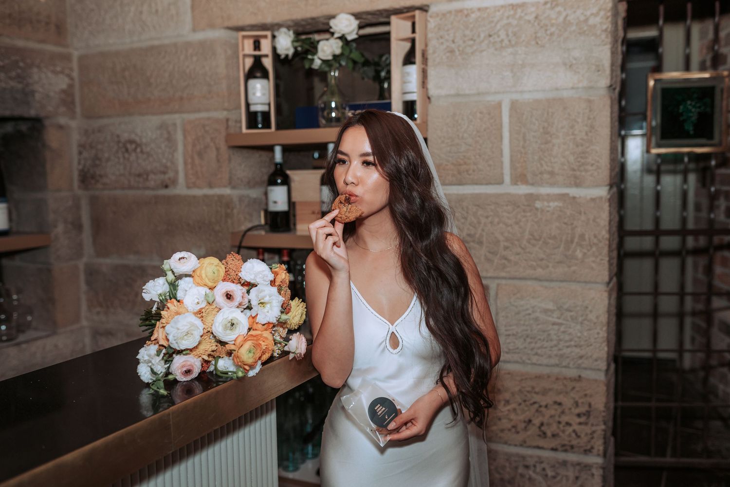 A bride in a white dress holds a beautiful bouquet while standing at a bar with stone walls.