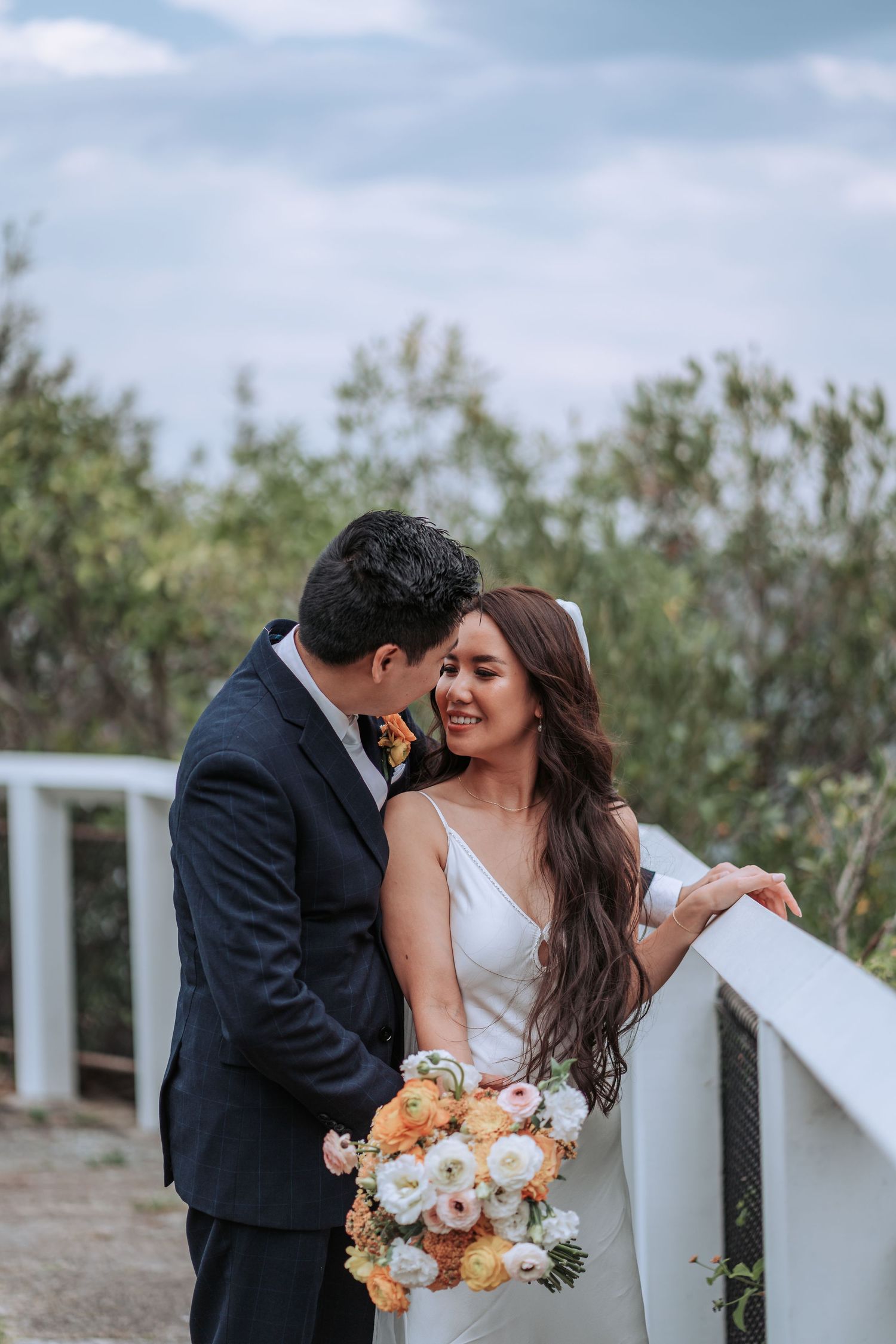 Romantic outdoor wedding portrait of couple by white railing with peach and white bouquet.