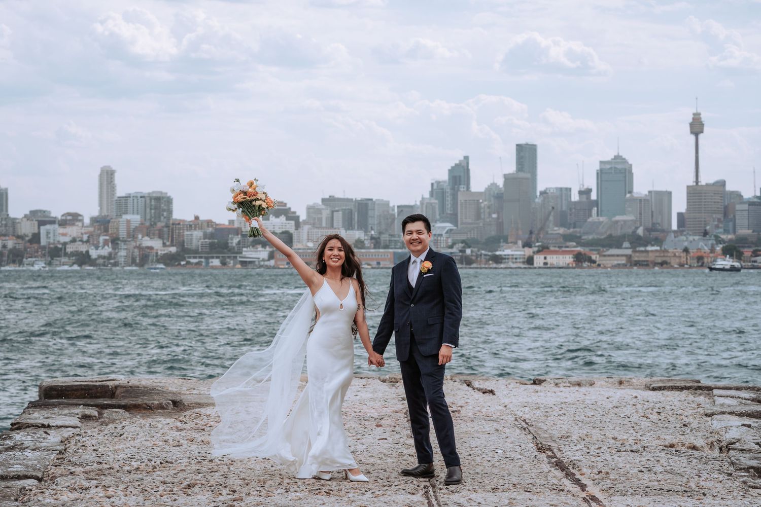 A wedding couple poses against the Sydney skyline backdrop while holding a bouquet.