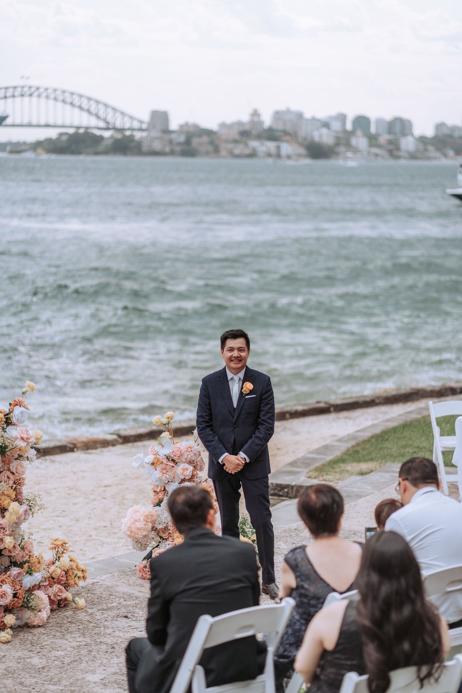 An outdoor wedding ceremony overlooking Sydney Harbour Bridge with florals and seated guests on a cloudy day.