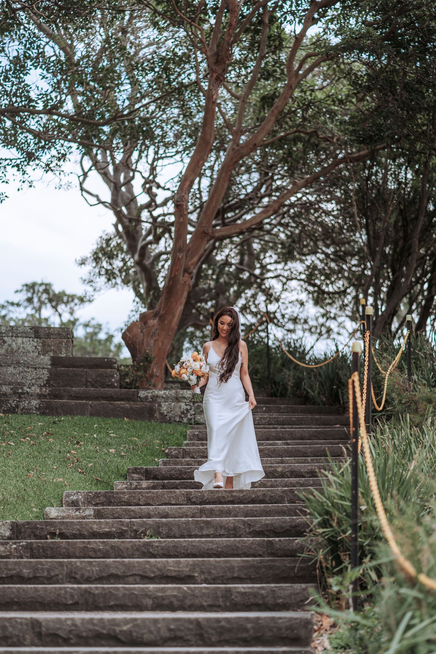 A bride in a flowing white dress descends stone steps surrounded by lush greenery and trees.