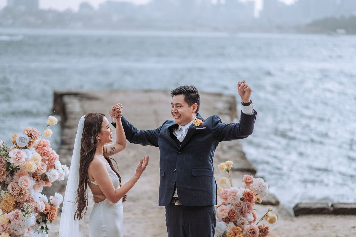 A joyful wedding celebration on a waterfront with peach floral arrangements and grey skies in the background.