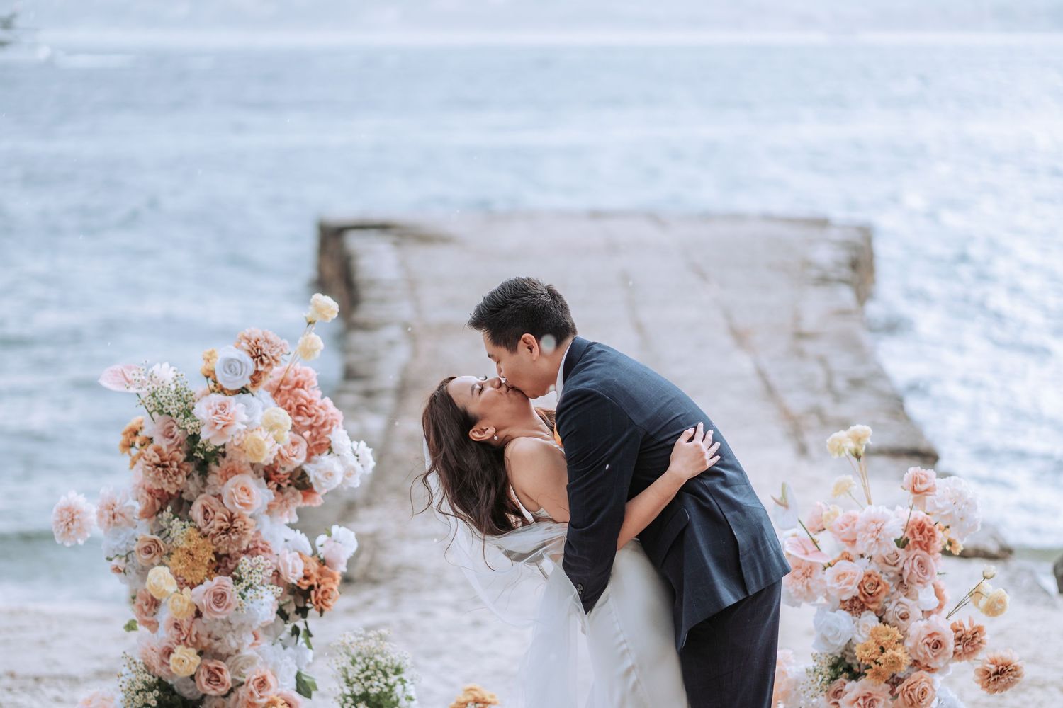 A romantic outdoor wedding moment on a pier with peach floral arrangements framing the couple.
