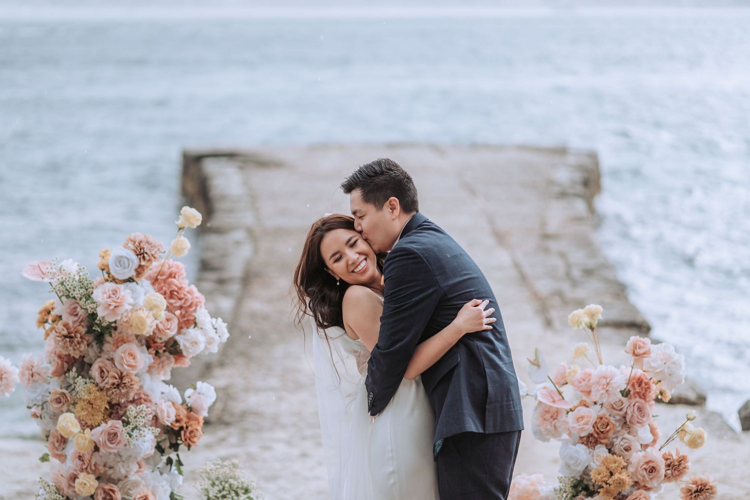 Romantic moment between bride and groom embracing on dock decorated with pink and peach floral arrangements.