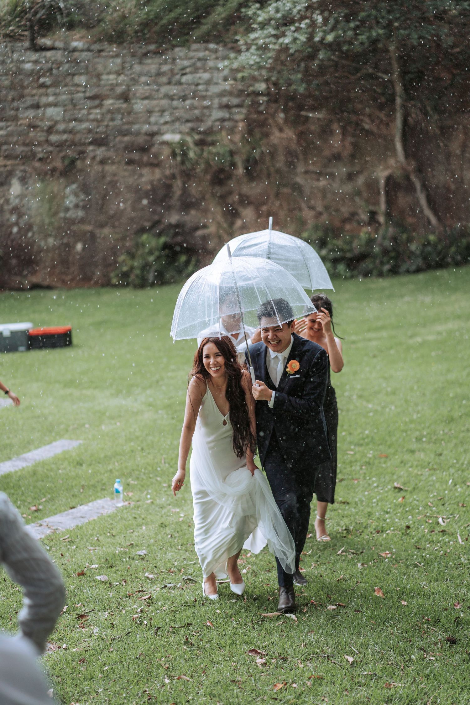 A couple walks under a clear umbrella in the rain during their wedding on a grassy hill.
