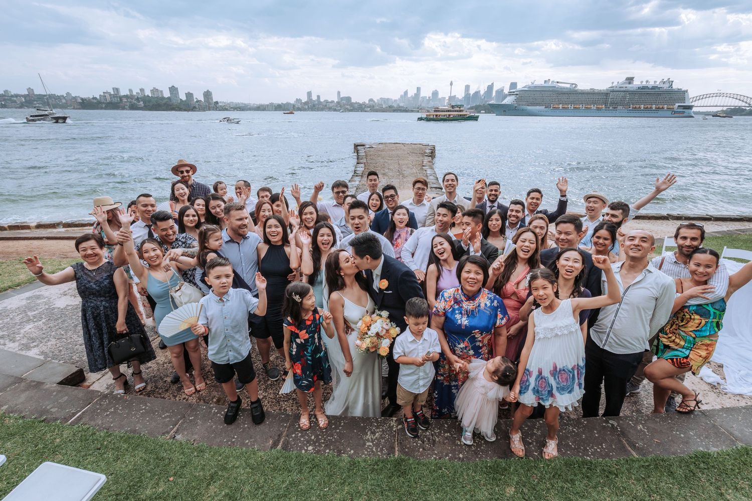 Large wedding party group photo taken at waterfront location with city skyline in background.
