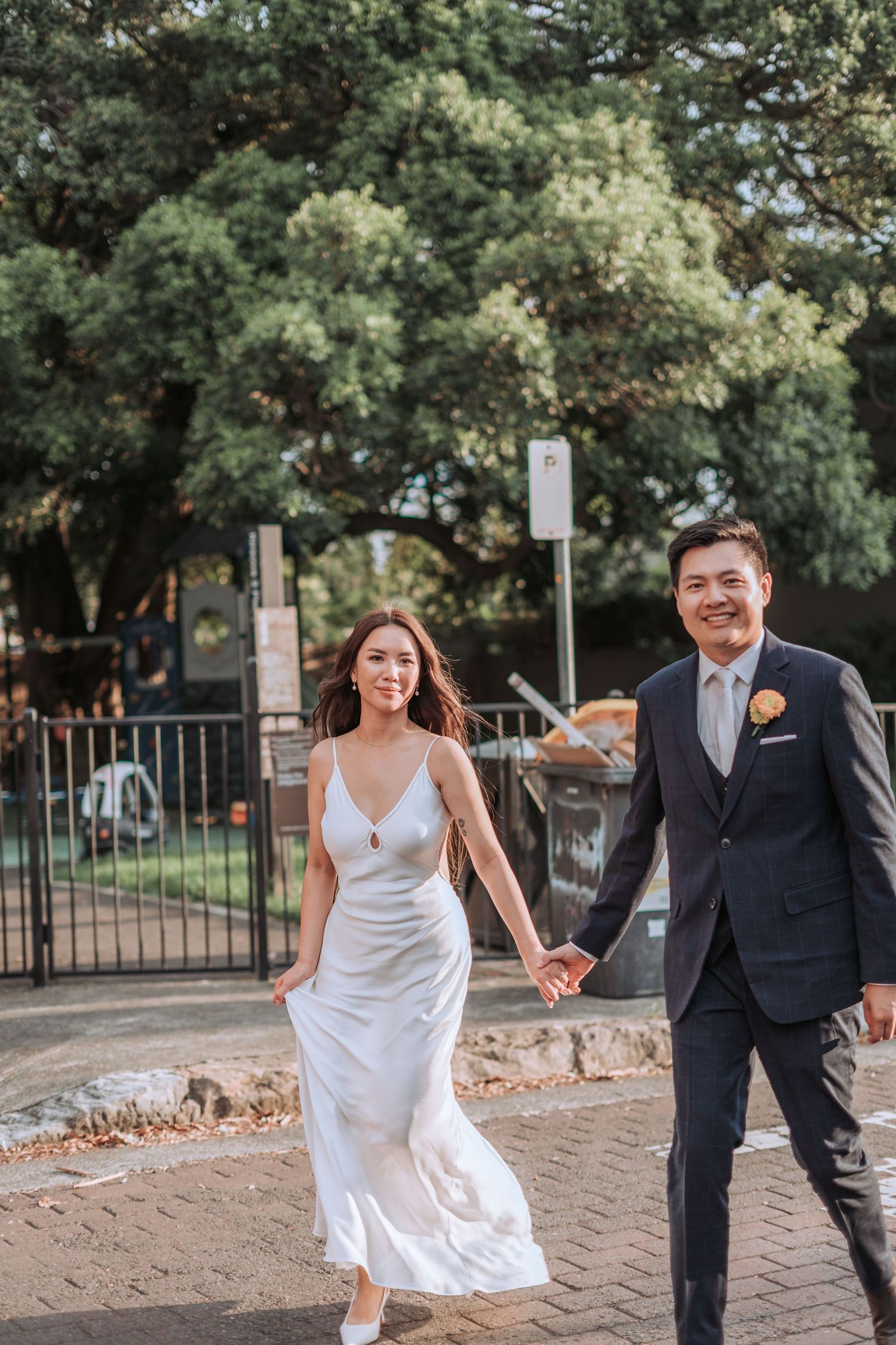 A newlywed couple walking together outdoors with lush green trees in the background.