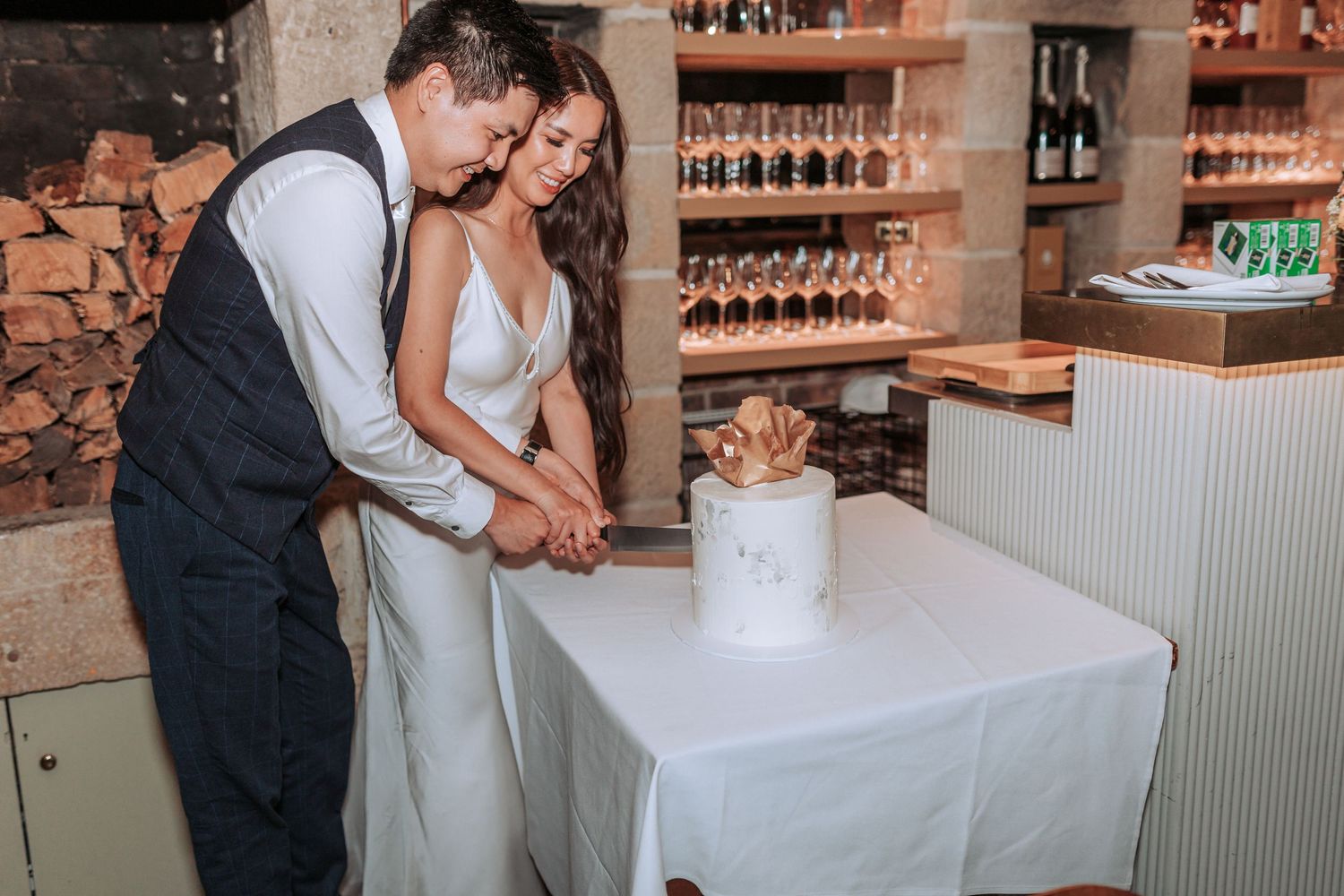 A couple cutting their wedding cake together at a rustic venue with exposed brick walls.
