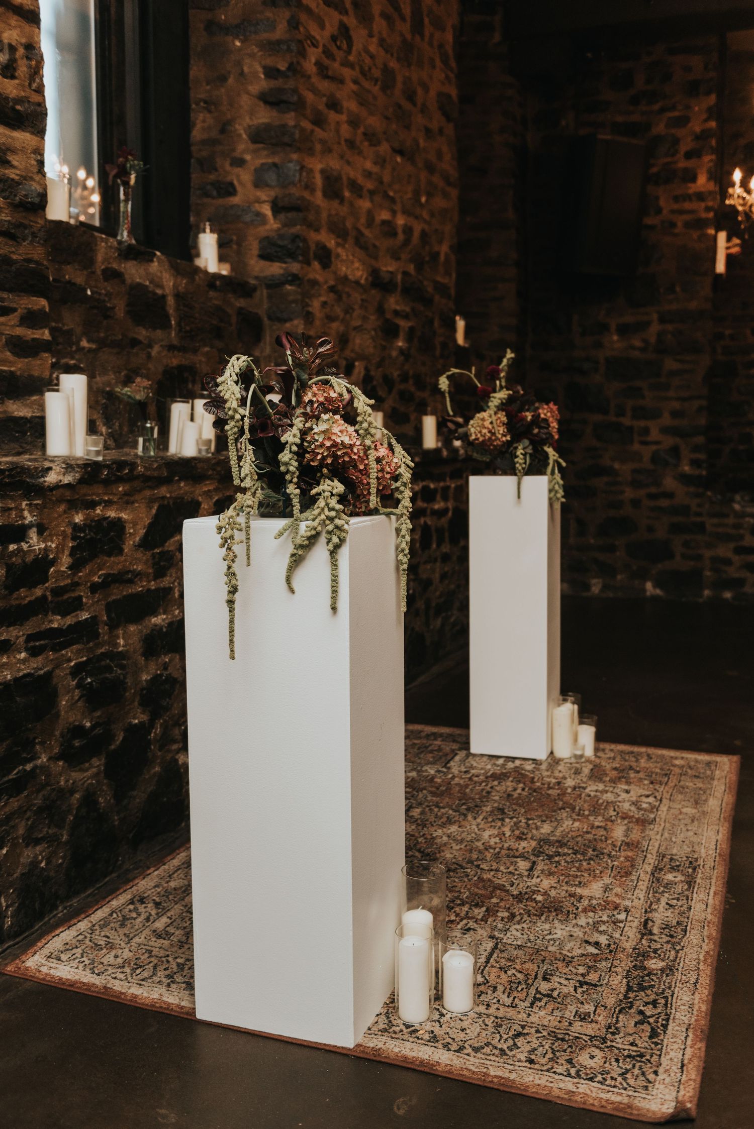 White pedestals with dried floral arrangements and candles create an elegant display against rustic stone walls.