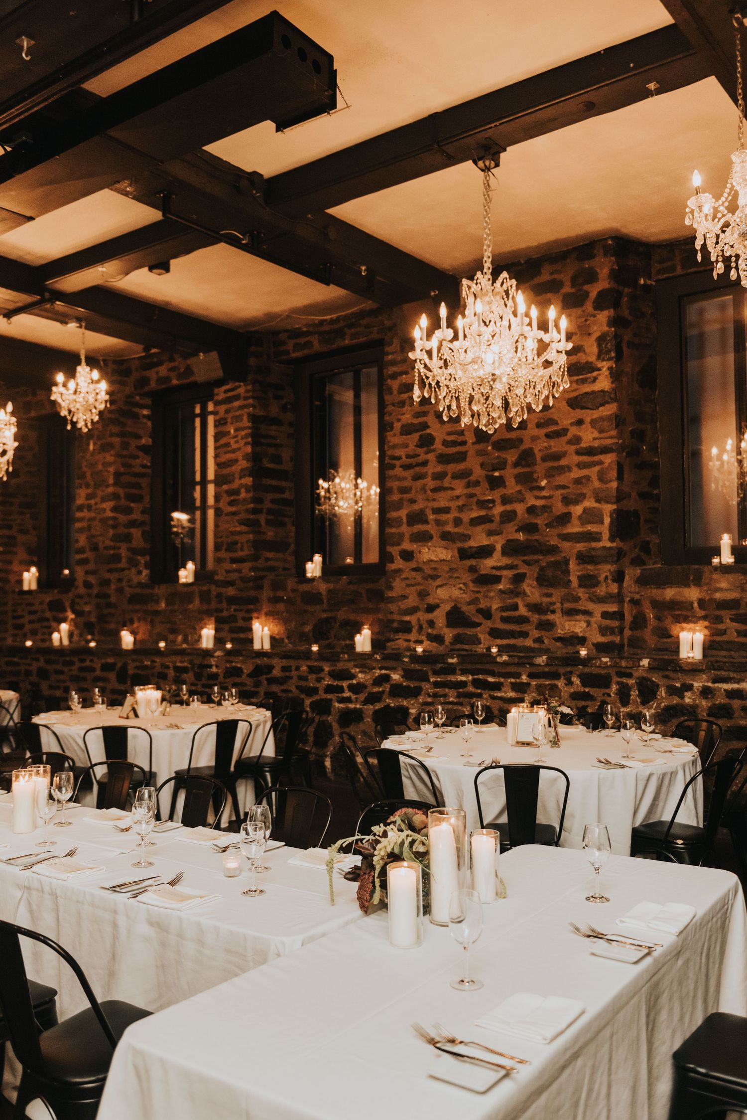 Rustic wedding reception venue with elegant crystal chandeliers, stone walls, and white tablecloths in warm ambient lighting.