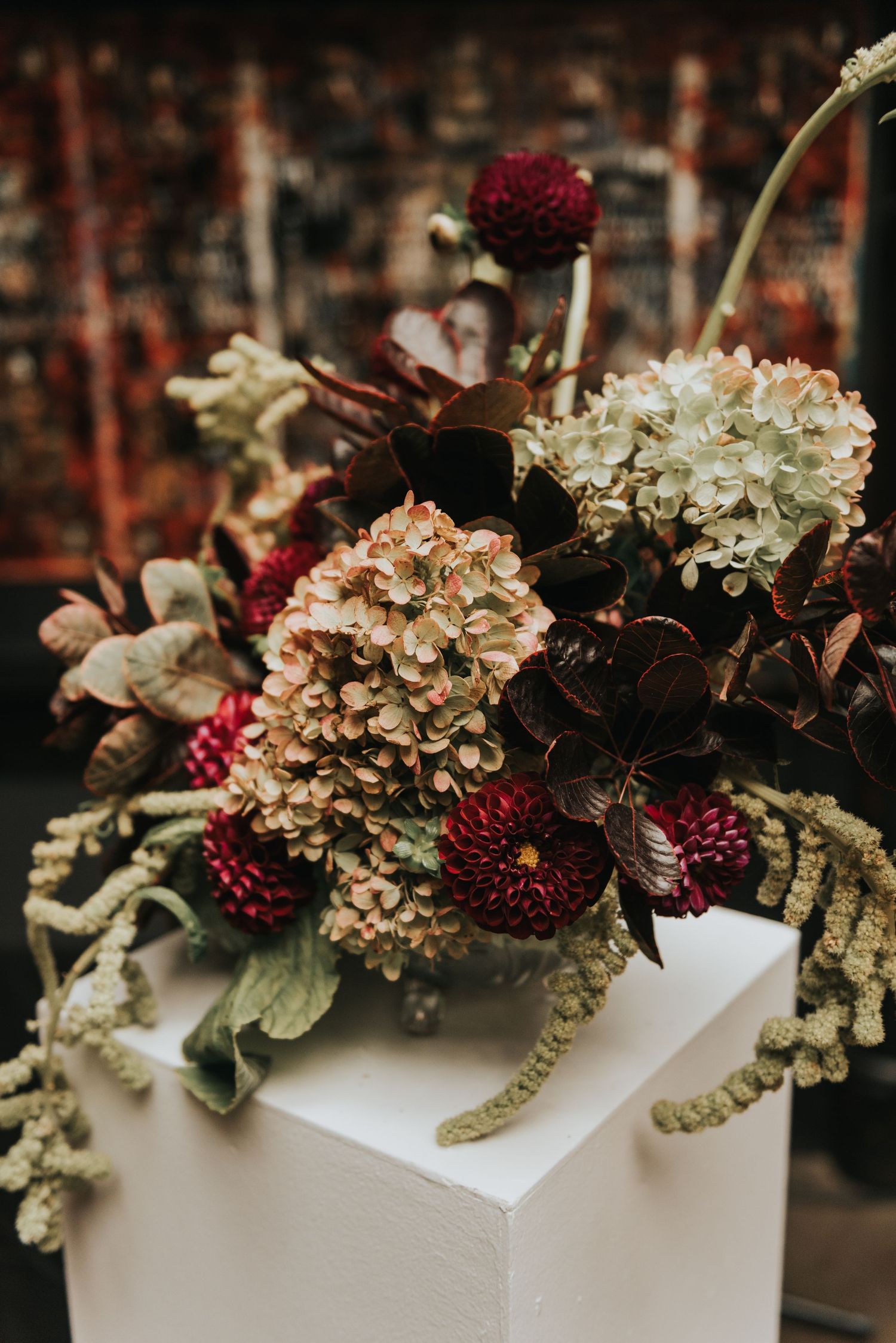 Moody fall flower arrangement with burgundy dahlias and dried hydrangeas on white pedestal in dark setting.