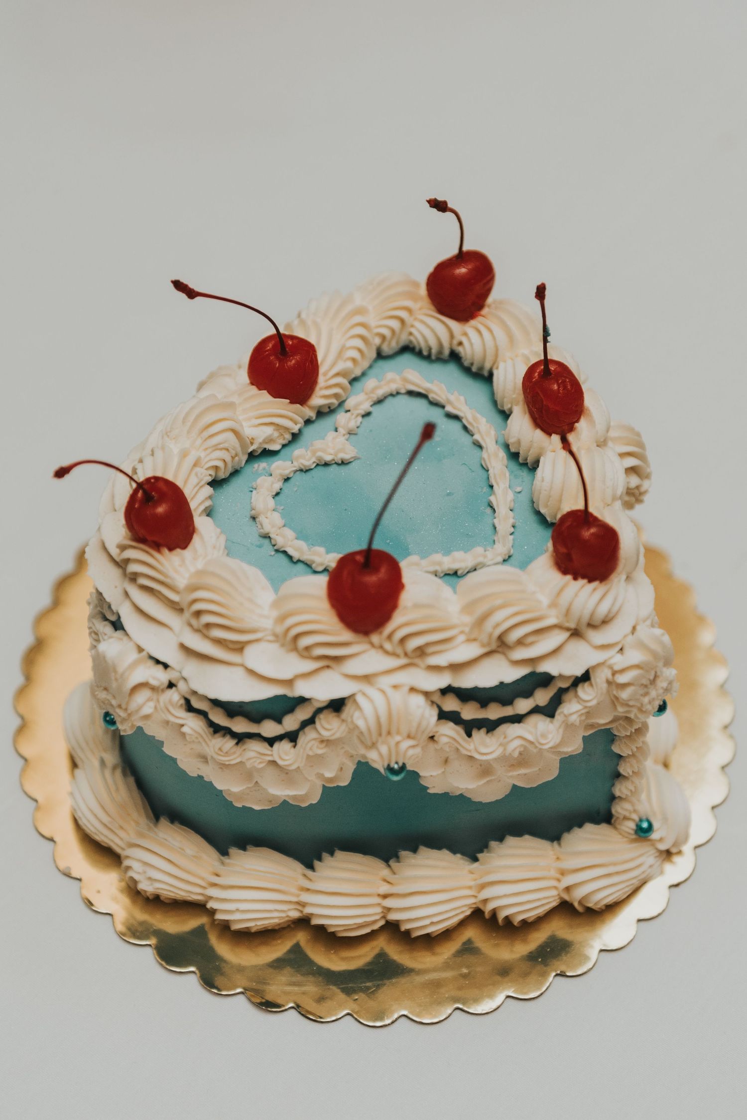A whimsical blue and white cake decorated with swirled frosting and topped with fresh red cherries on a golden cardboard base.
