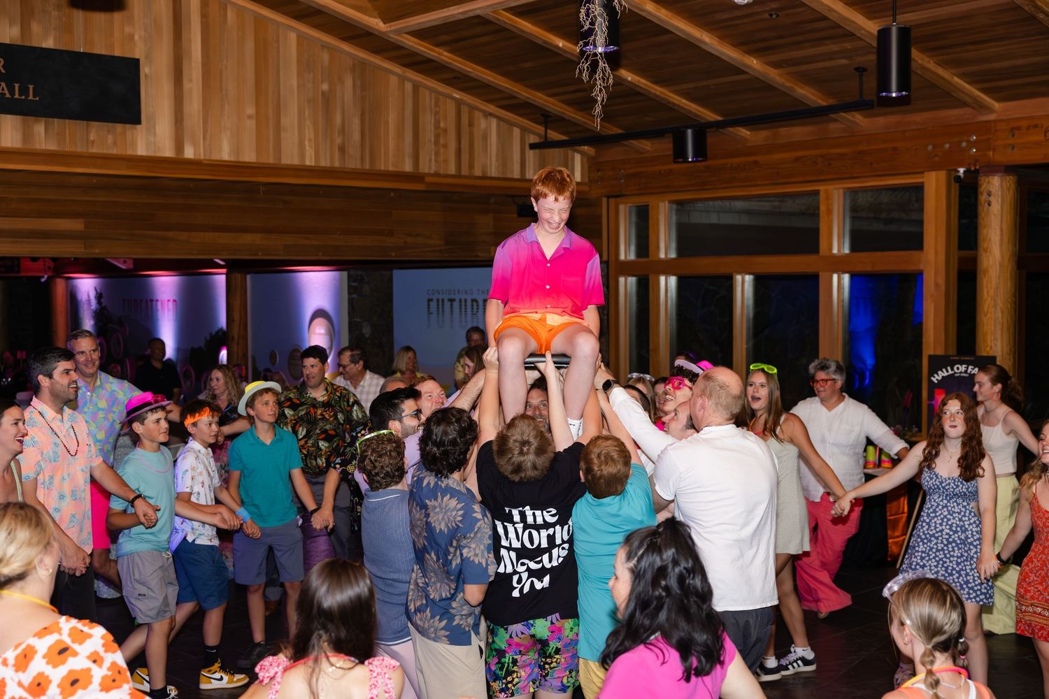 A celebratory crowd lifts someone wearing a bright pink shirt during a lively party in a wood-paneled venue.