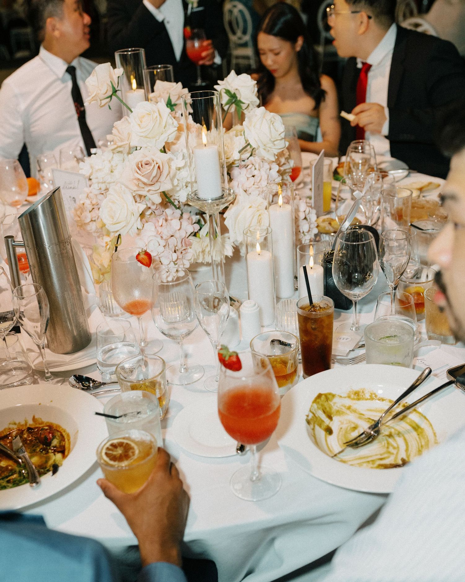 Elegant wedding reception table set with glasses, drinks, and plates of food amidst soft candlelight and floral decorations.