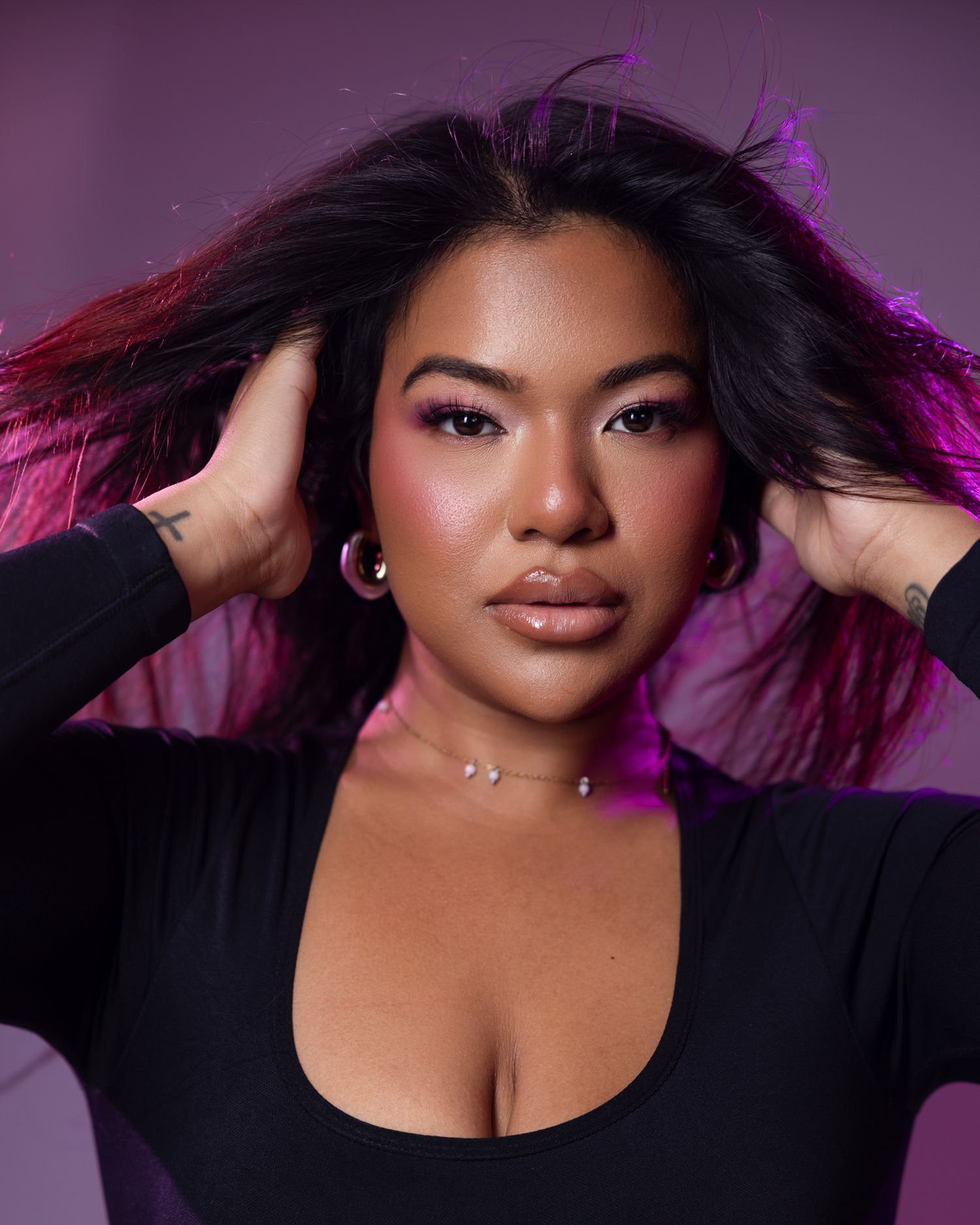 Model with flowing dark hair in black top poses against purple backdrop with dramatic pink lighting.