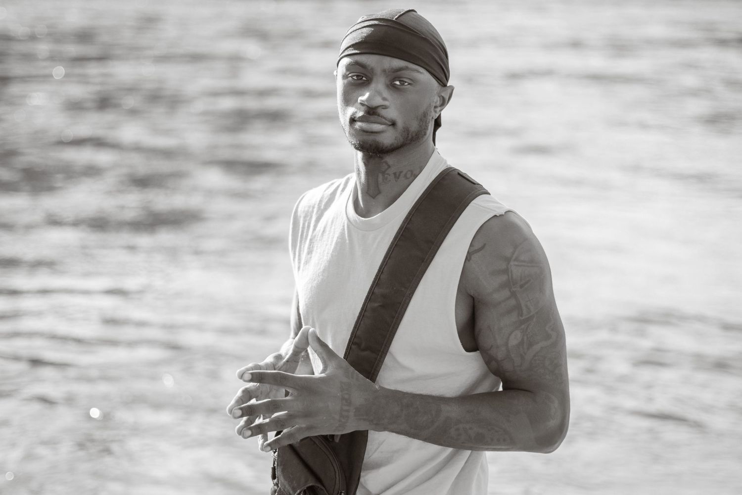 A person in a tank top and bandana stands thoughtfully against a water backdrop in black and white.