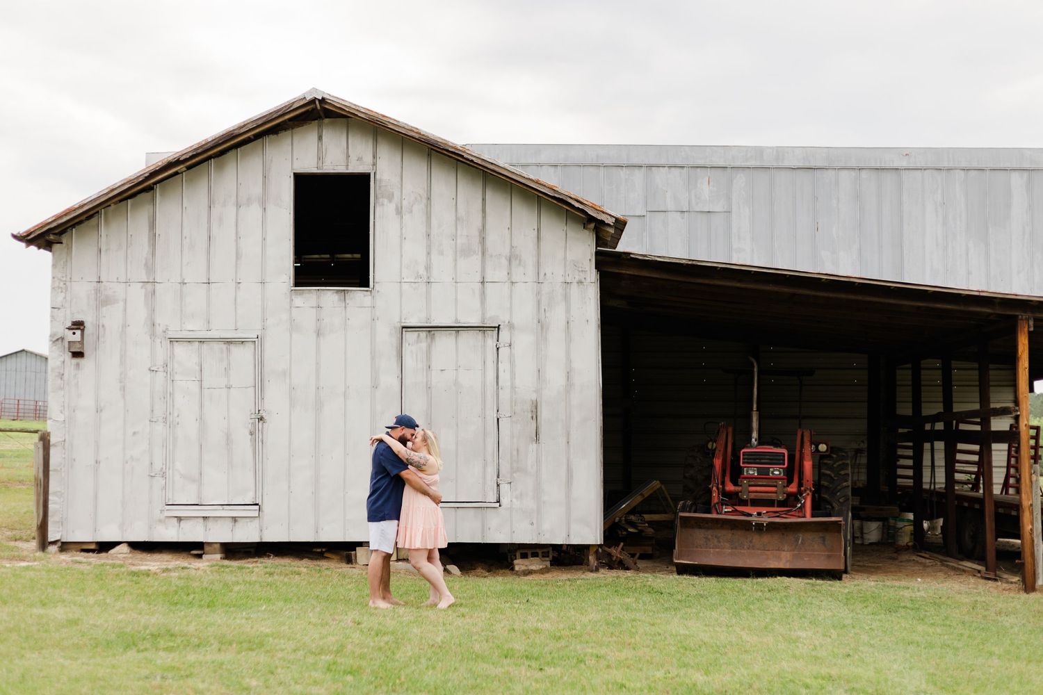Romantic couple embracing in front of rustic white barn with vintage farm equipment.