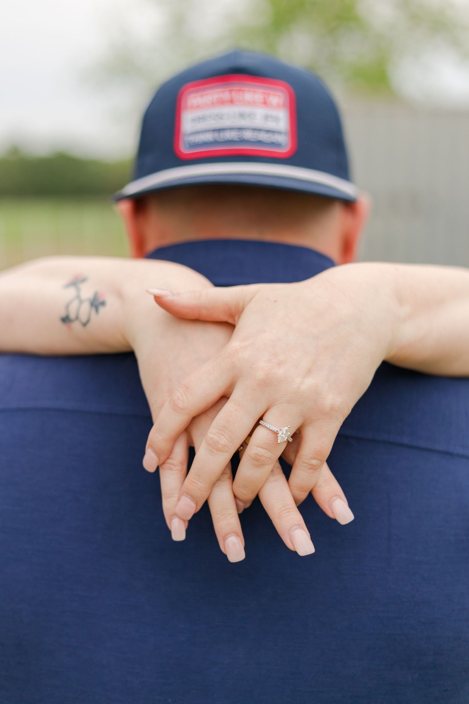 Close-up of hands wearing engagement ring with baseball cap and tattoo detail.