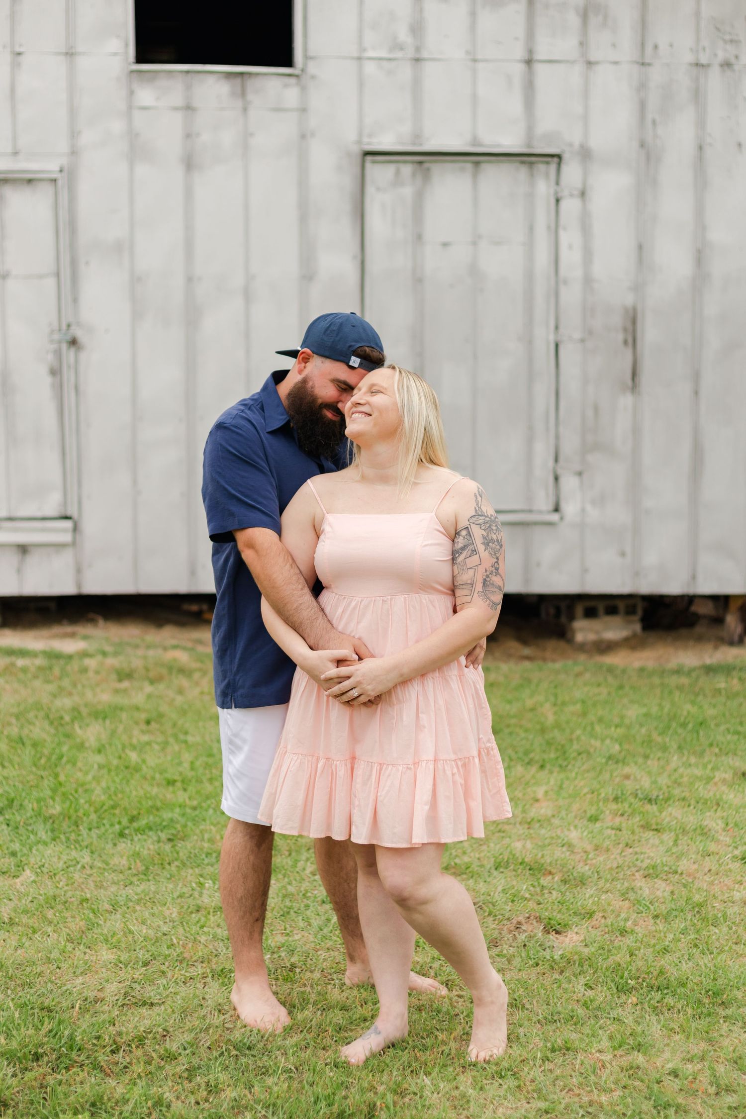 Couple in casual attire share tender moment outside white wooden barn.