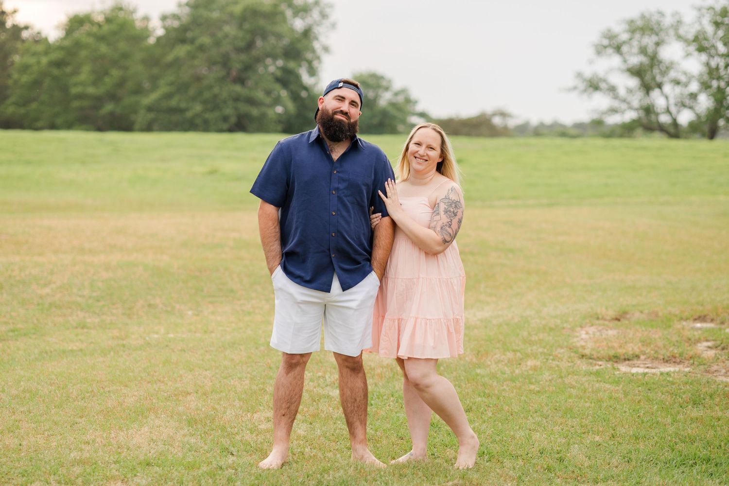 A couple poses casually together in a grassy field wearing blue and pink outfits.