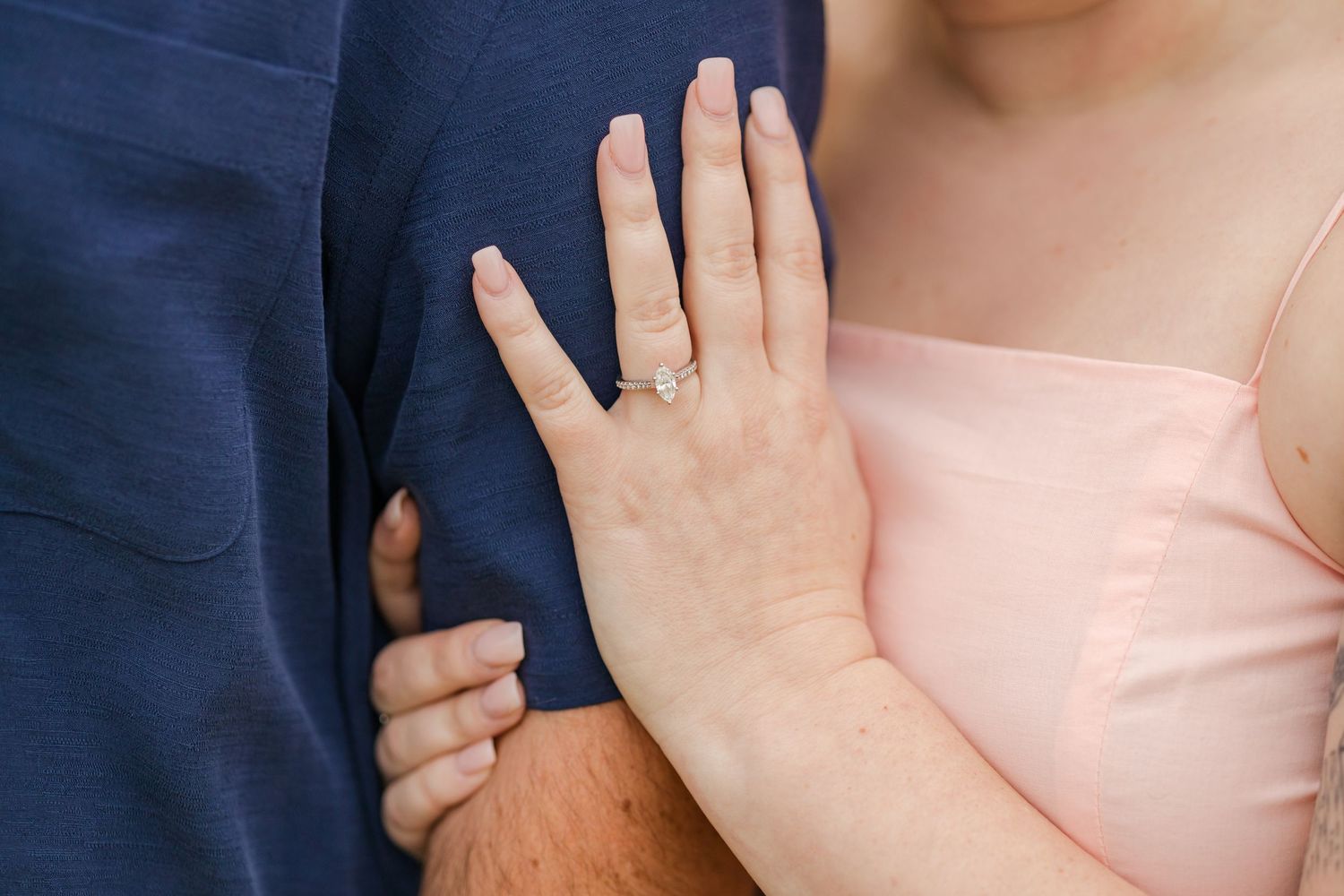A delicate engagement ring sparkles as a hand rests against a navy blue shirt.