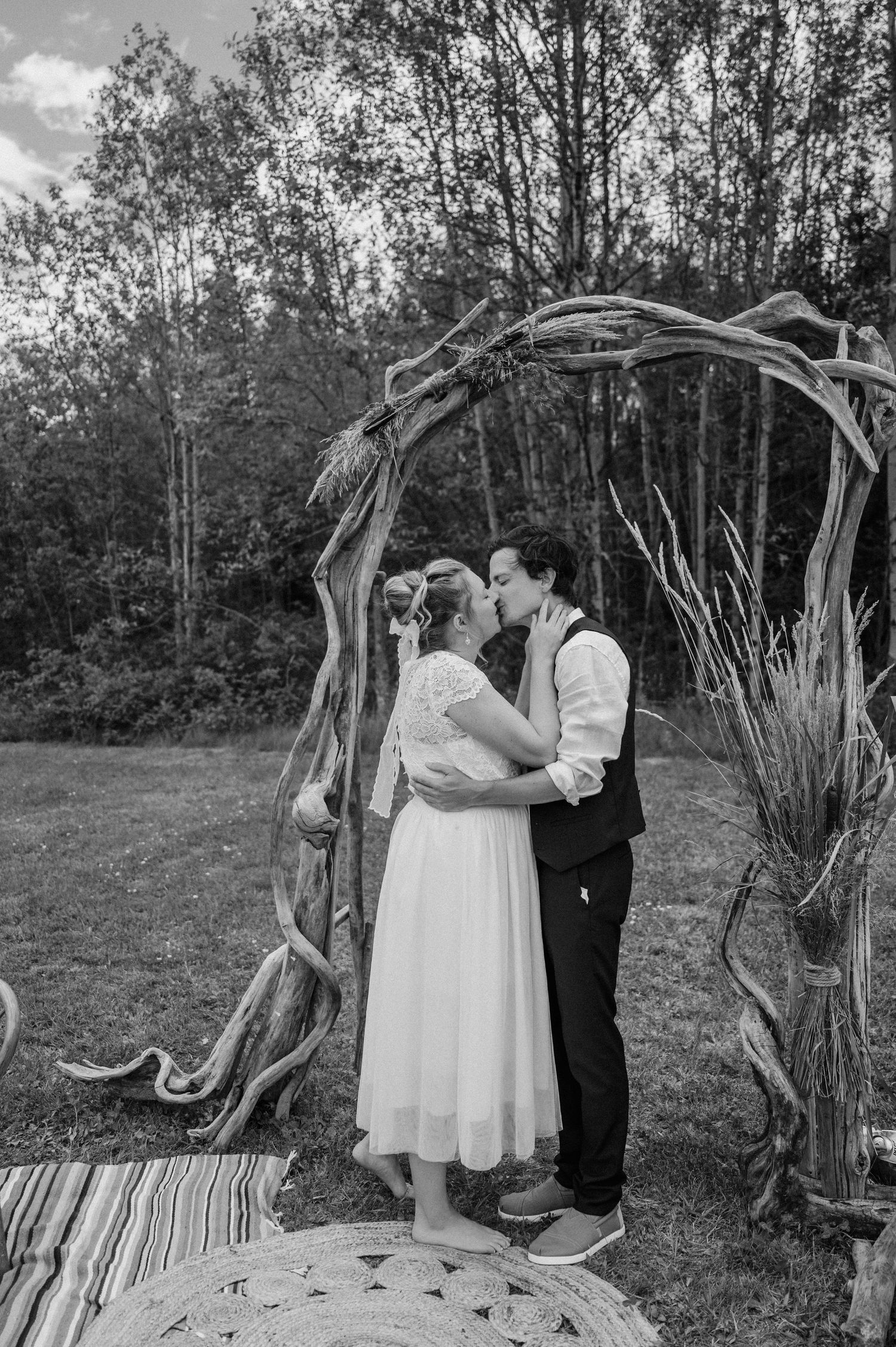 A romantic black and white wedding photo under a rustic driftwood arch in a forest setting.