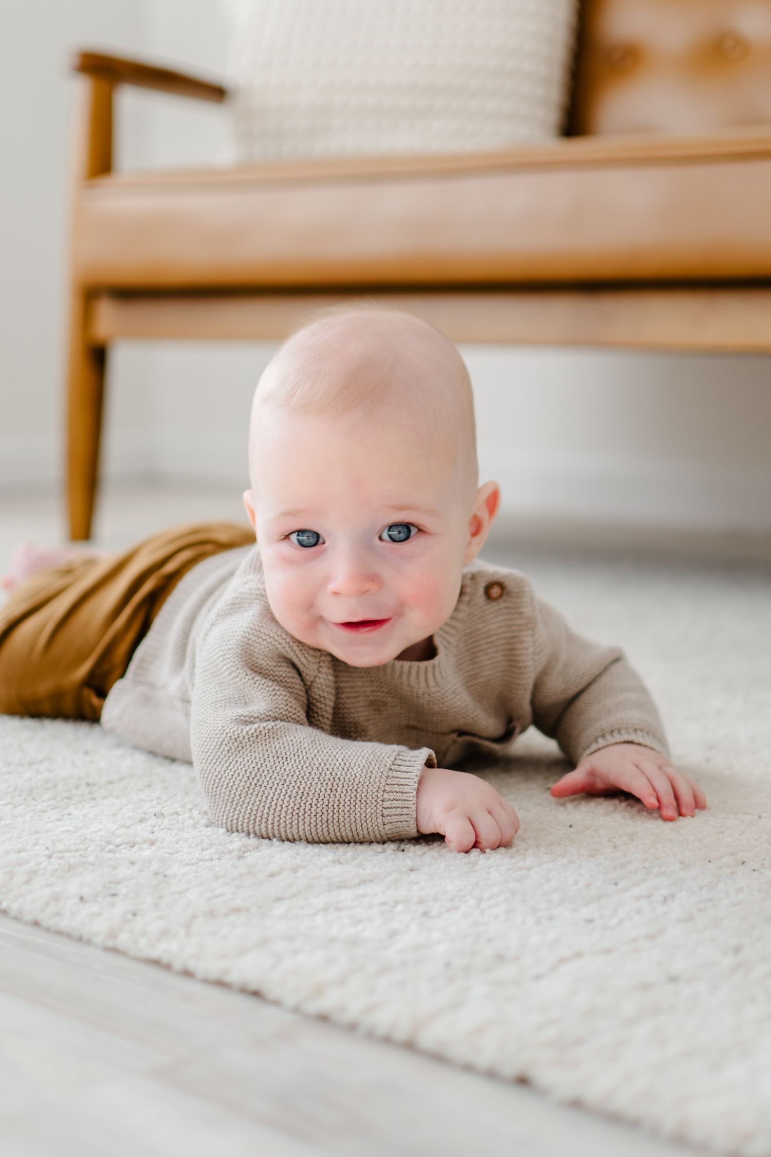 Adorable baby wearing a beige sweater lies on plush white carpet showing a sweet smile.