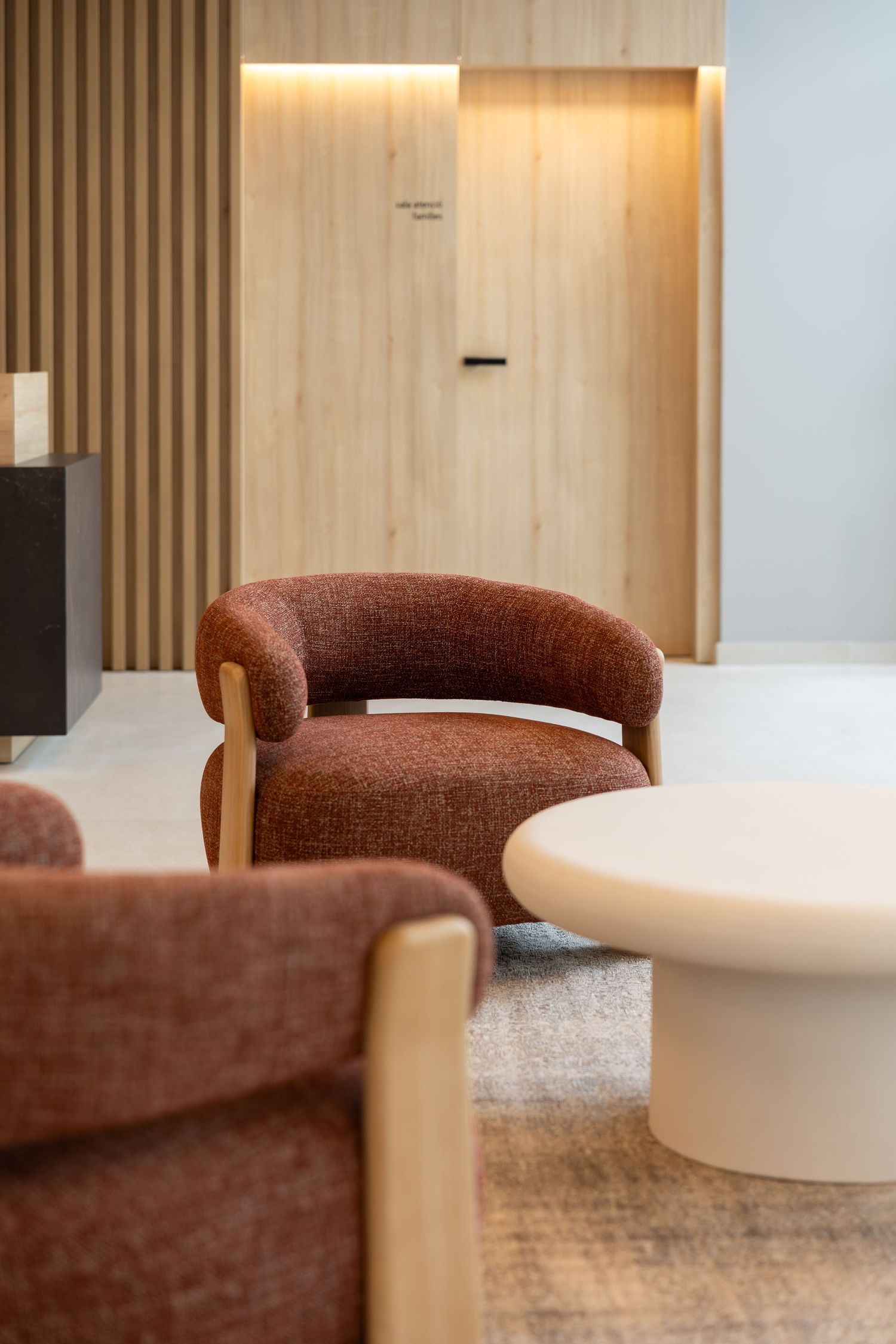 Modern lounge area featuring rust-colored curved armchairs with wood frames and a white round coffee table against warm wood paneling.