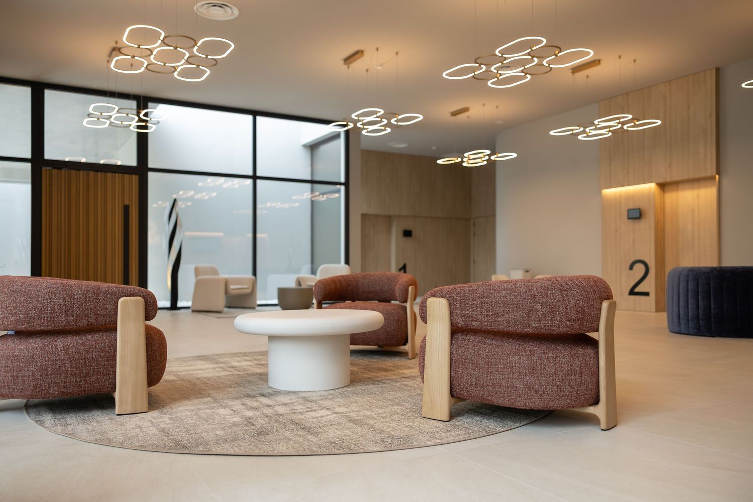 Modern hotel lobby with circular pendant lights, plush mauve armchairs, a white round coffee table, and floor-to-ceiling windows.