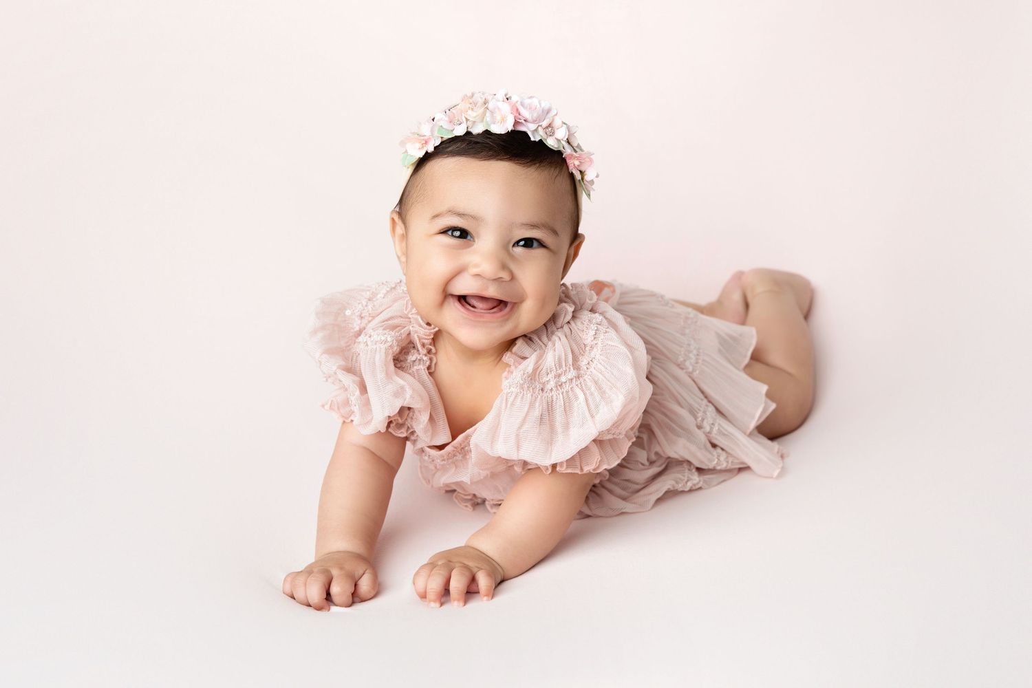 Smiling baby girl in pink floral romper and flower crown doing tummy time on white background.