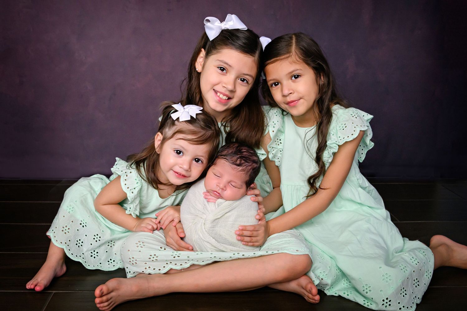Three older sisters in mint dresses lovingly hold their newborn sibling in a professional portrait photo. Photographer in Cypress TX