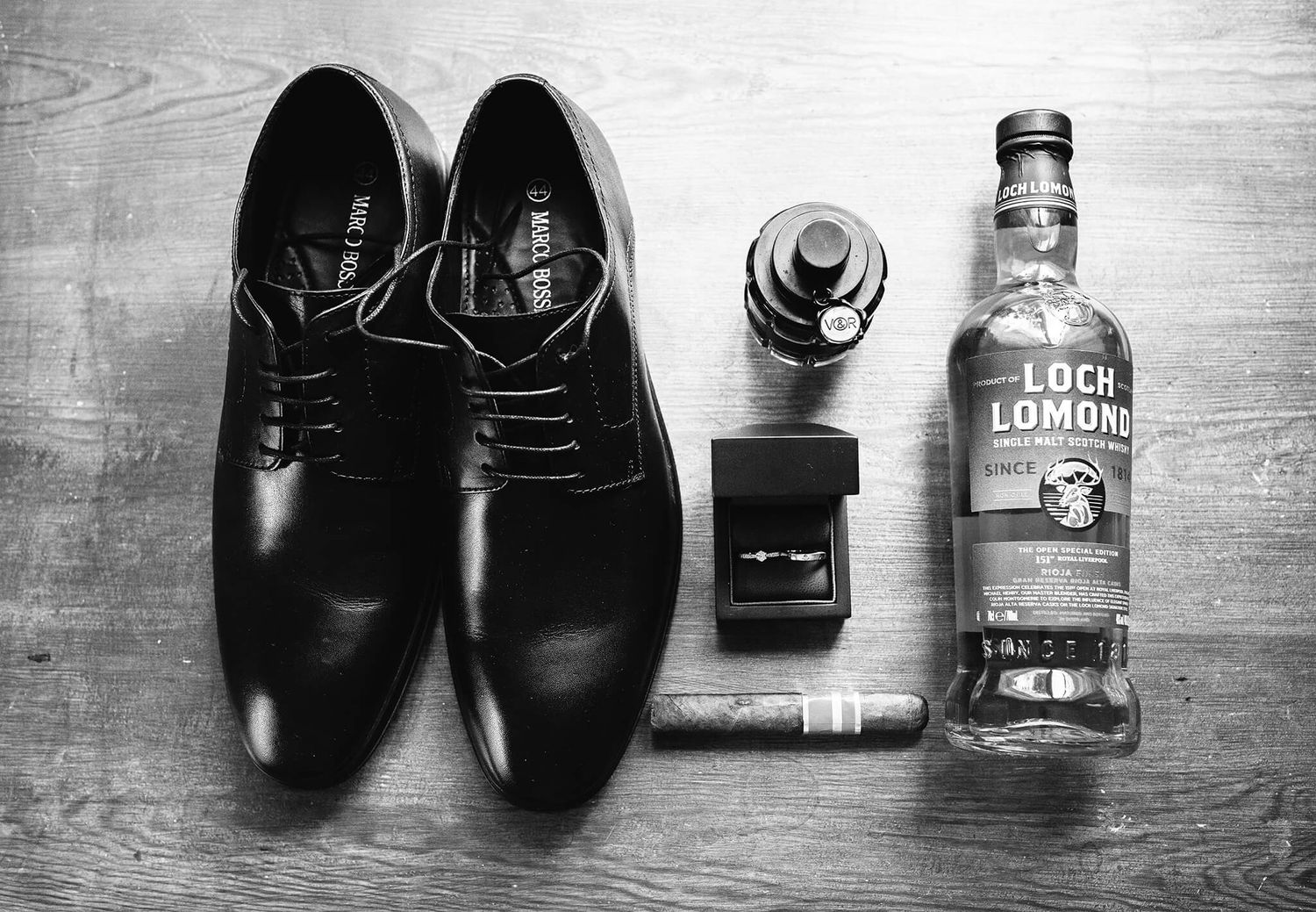 Black and white flat lay of groom accessories including dress shoes, whiskey bottle, cigar and ring box.