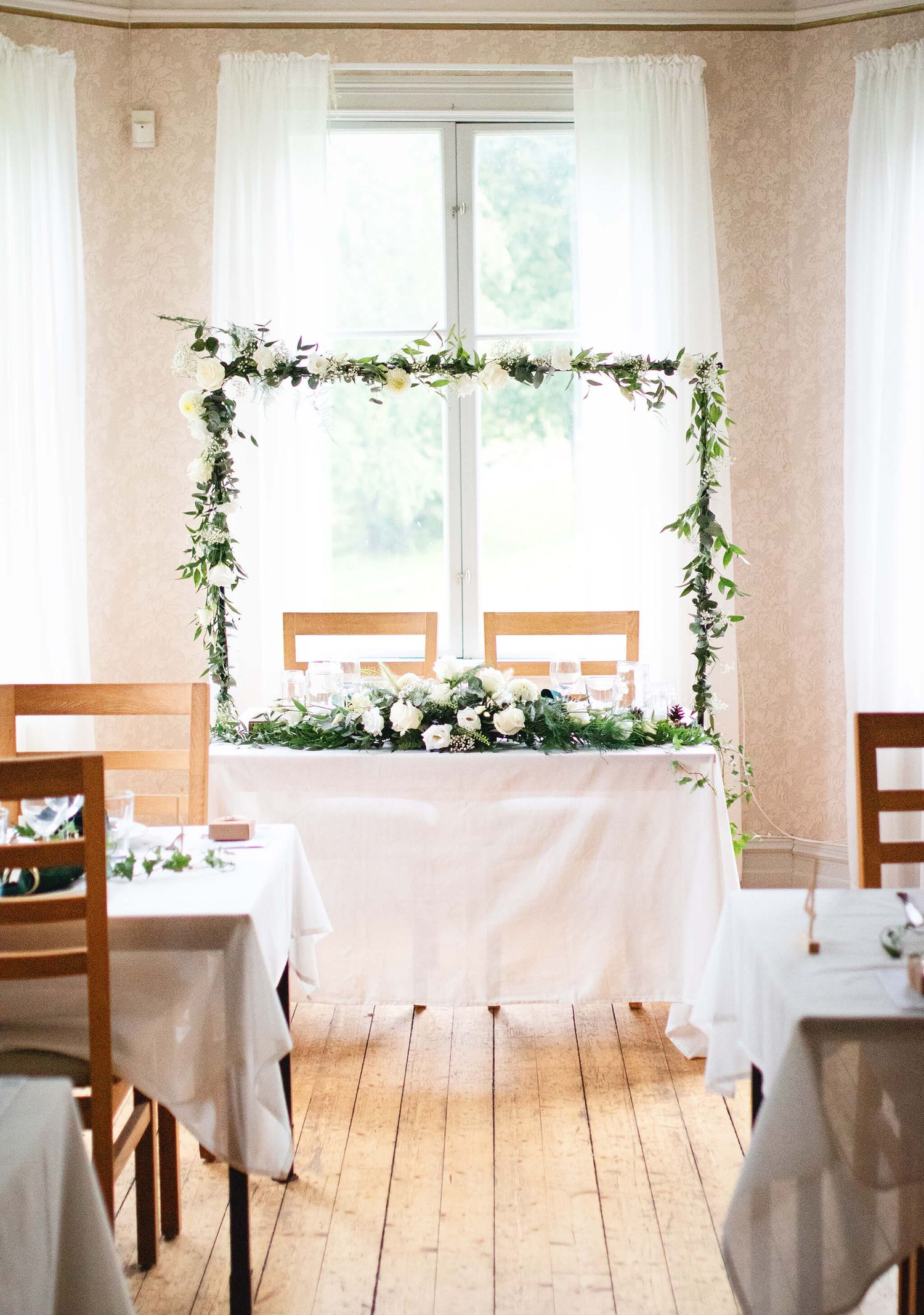 Elegant wedding reception sweetheart table decorated with a floral arch and greenery garland by a bright window.