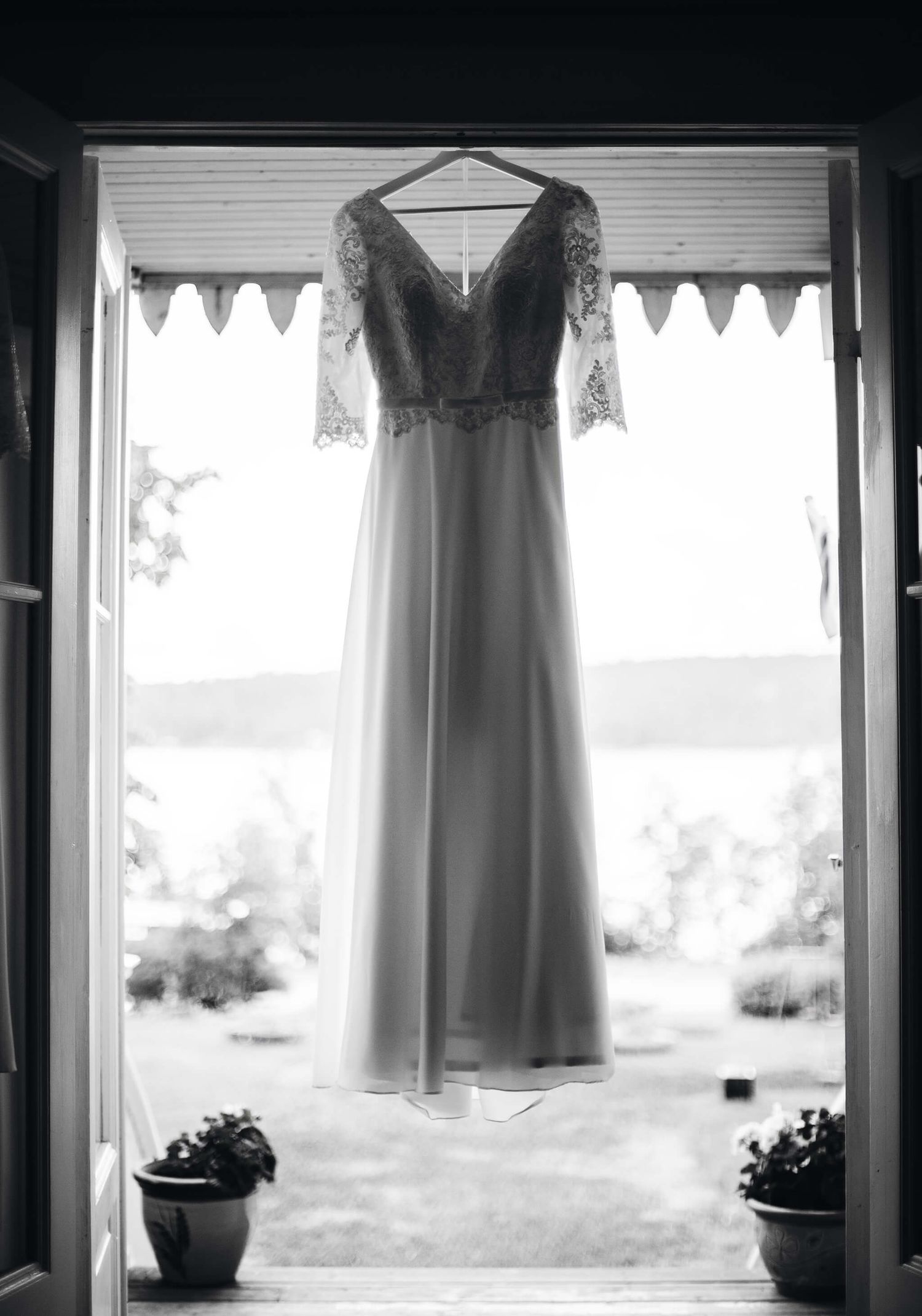 Black and white photo of a lace wedding dress hanging in a doorway with a scenic water view behind.