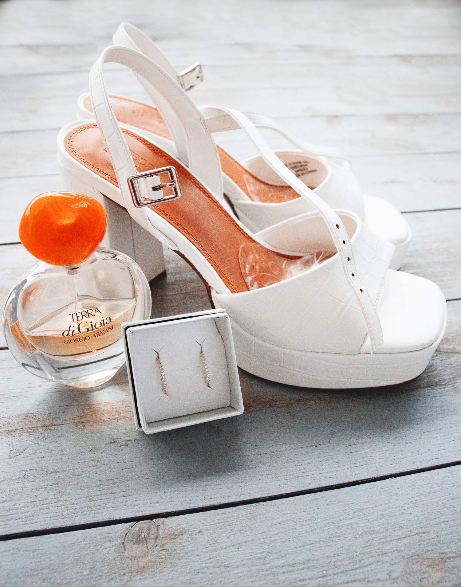 White bridal heels with orange interior displayed beside perfume bottle and earrings on rustic wooden surface.