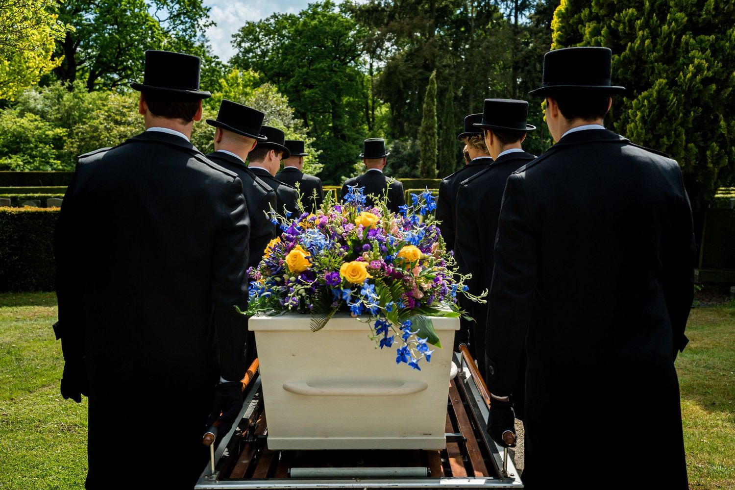 Funeral pallbearers in black top hats carry a white casket adorned with colorful flowers through a green cemetery.