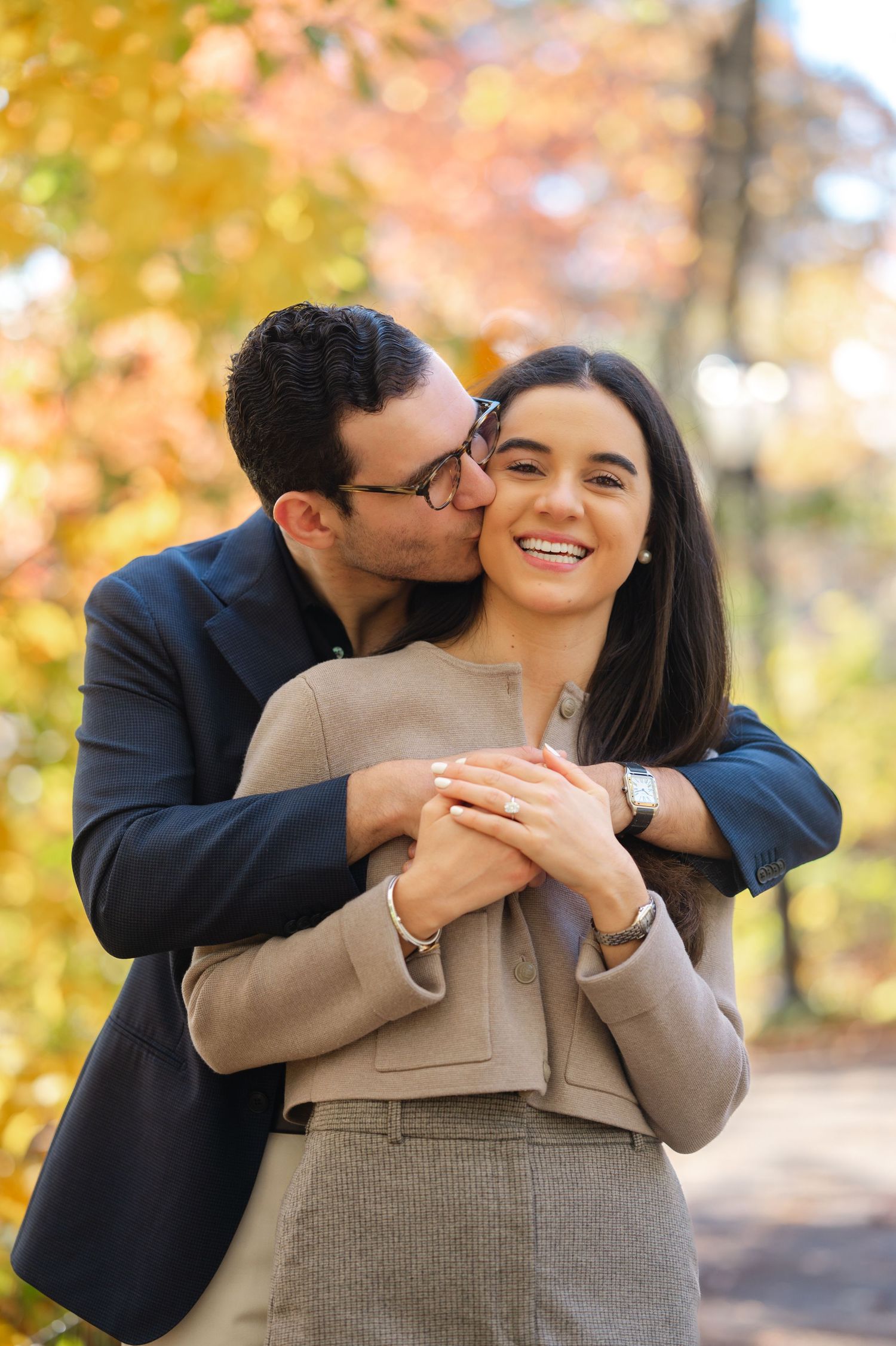 A couple embraces in an autumn park, man kissing woman on cheek while she smiles, wearing a ring after their proposal in Central Park.