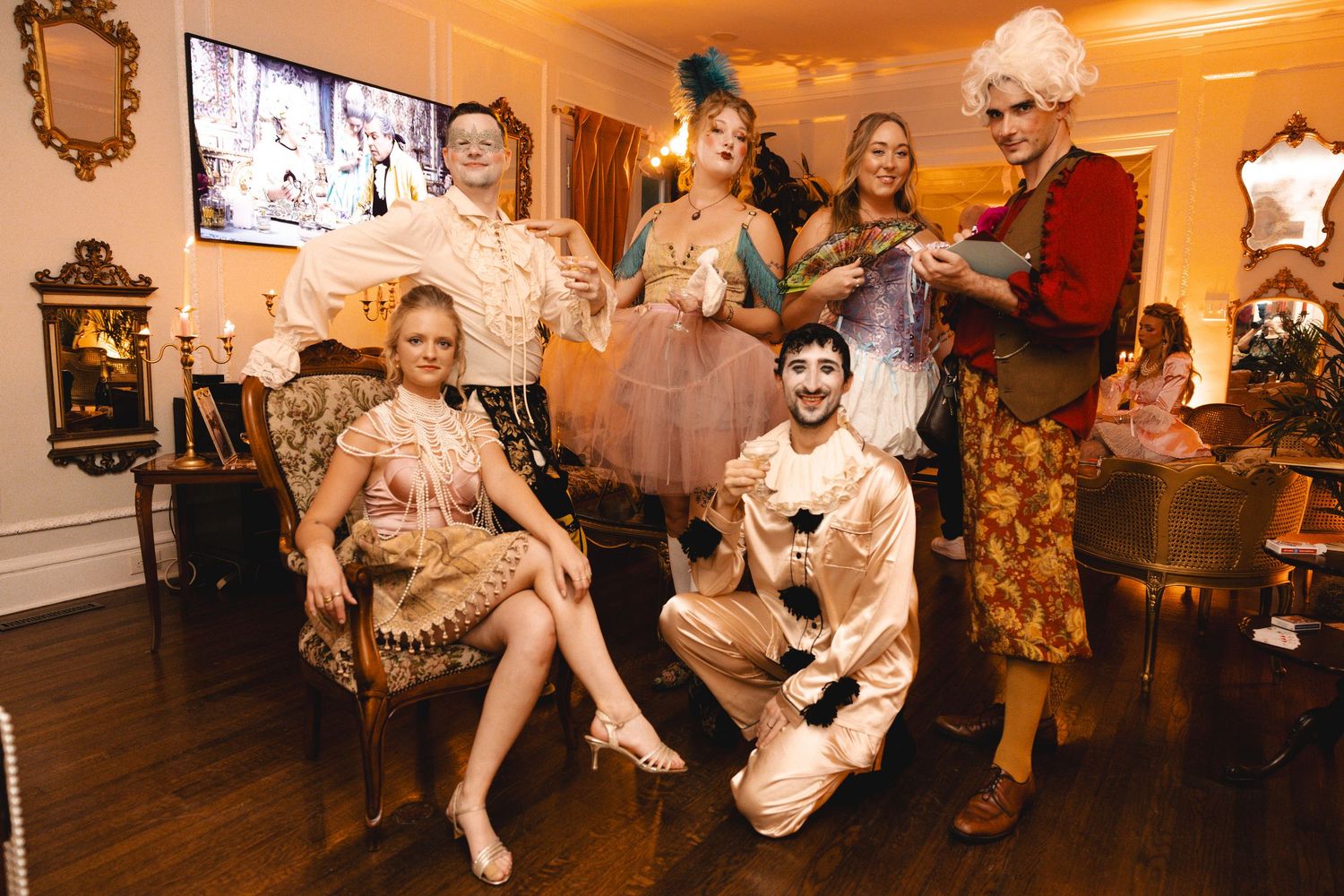 Group of six people in colorful vintage costumes posing together at an elegant indoor party event.