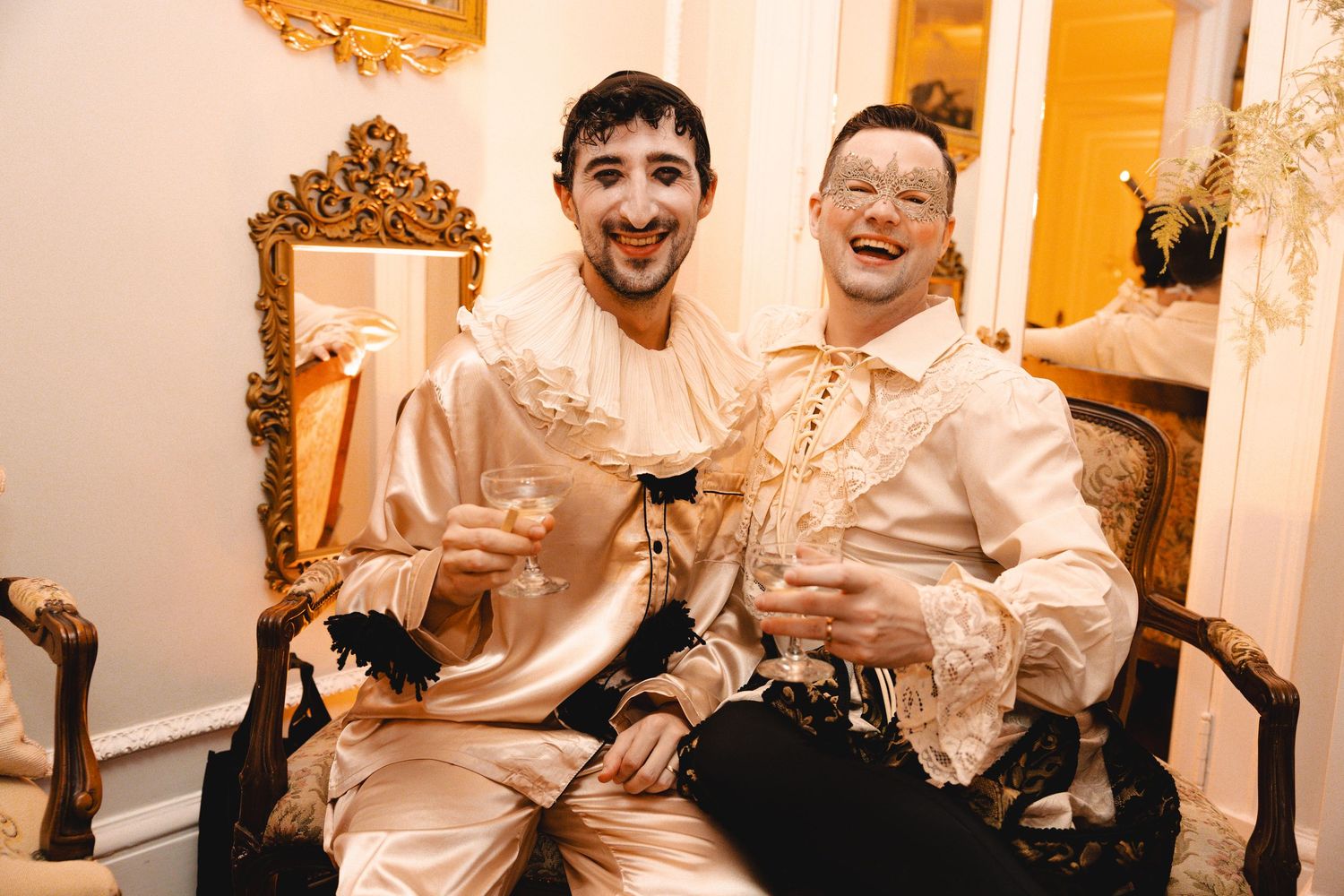 Two men in elegant white masquerade costumes with gold masks laugh together at a fancy costume party.